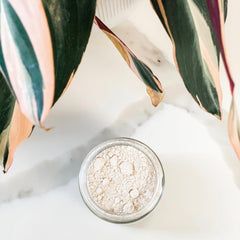 Powder in glass jar with light beige color, marble background, and green and pink plant leaves