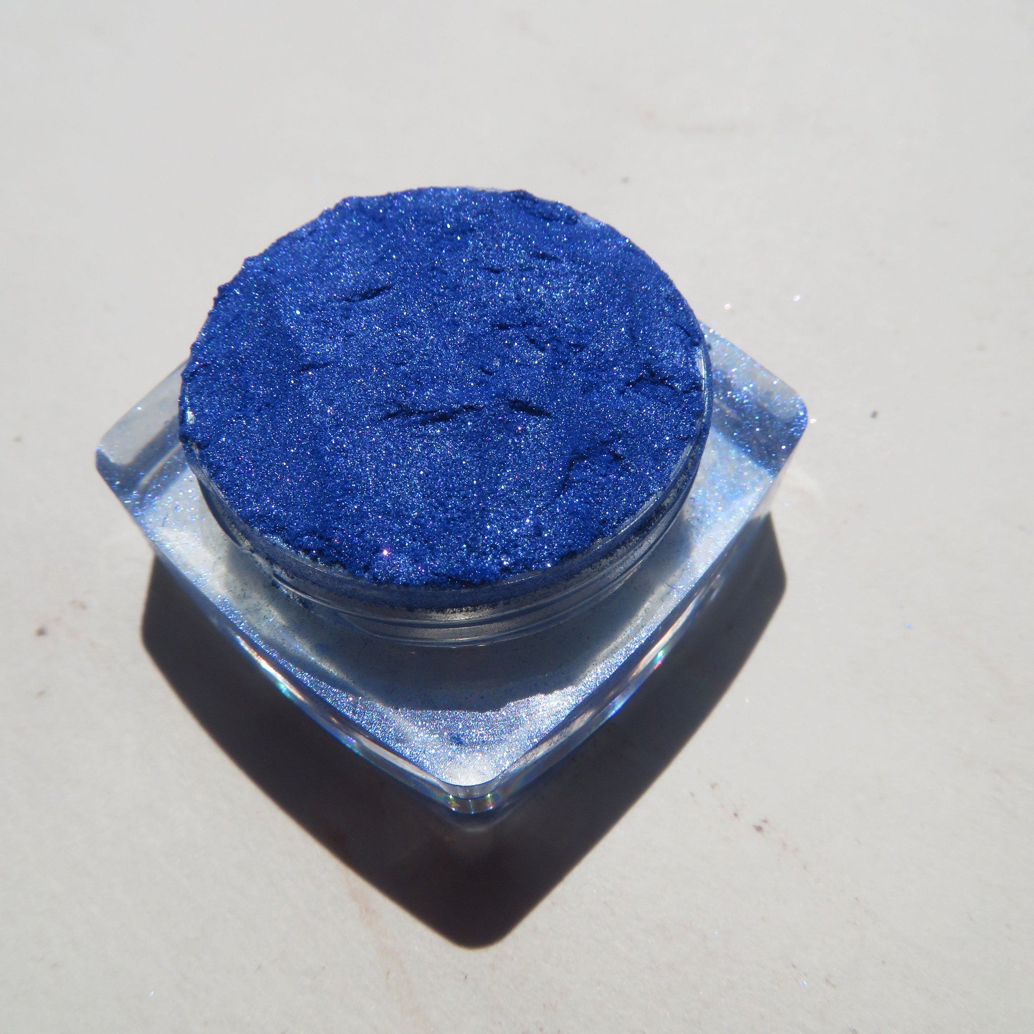 Close-up of vibrant blue shimmer loose mineral eyeshadow with reflective particles in a small glass jar