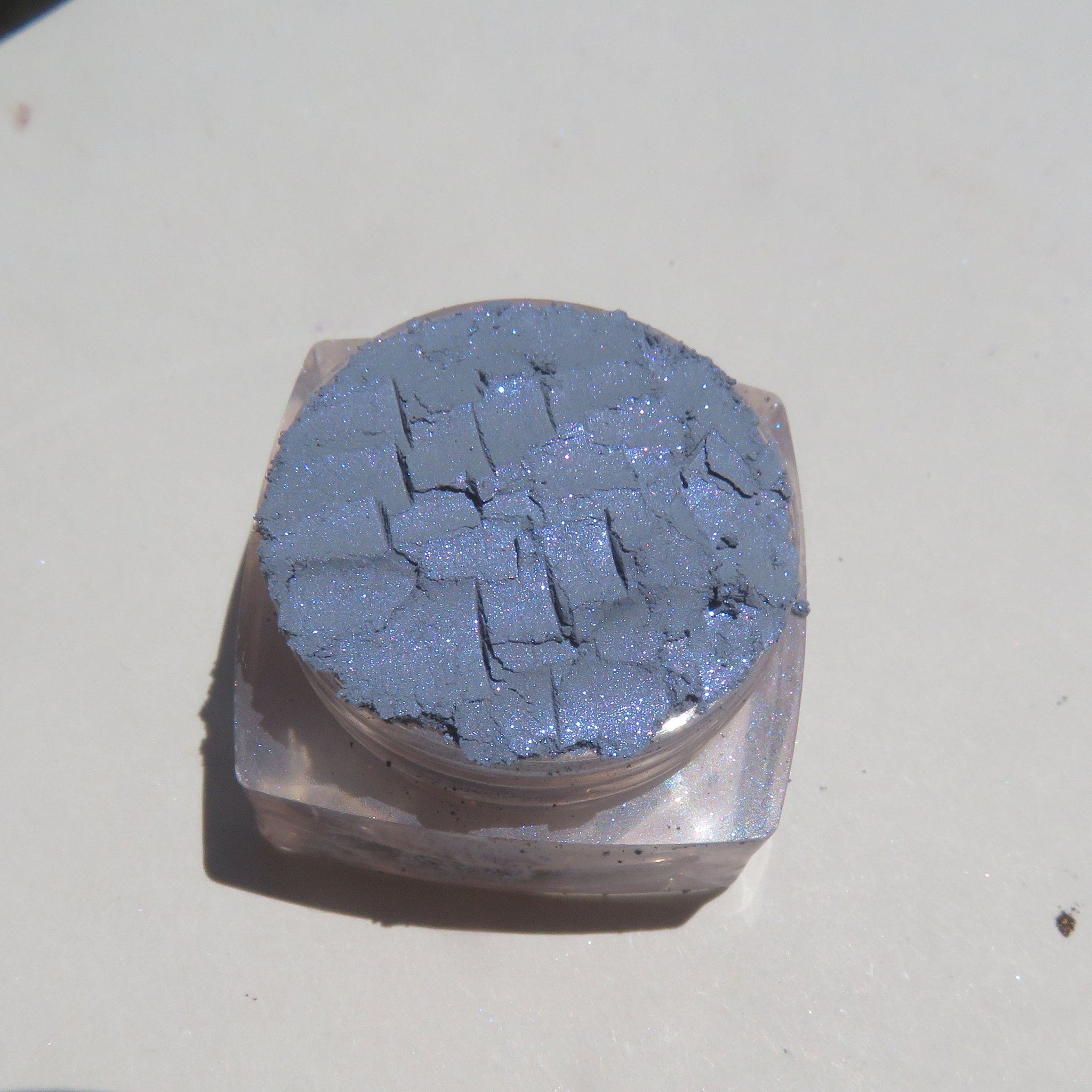 Close-up of loose Enigma Blue Shimmer mineral eyeshadow with reflective particles in vegan, cruelty-free packaging