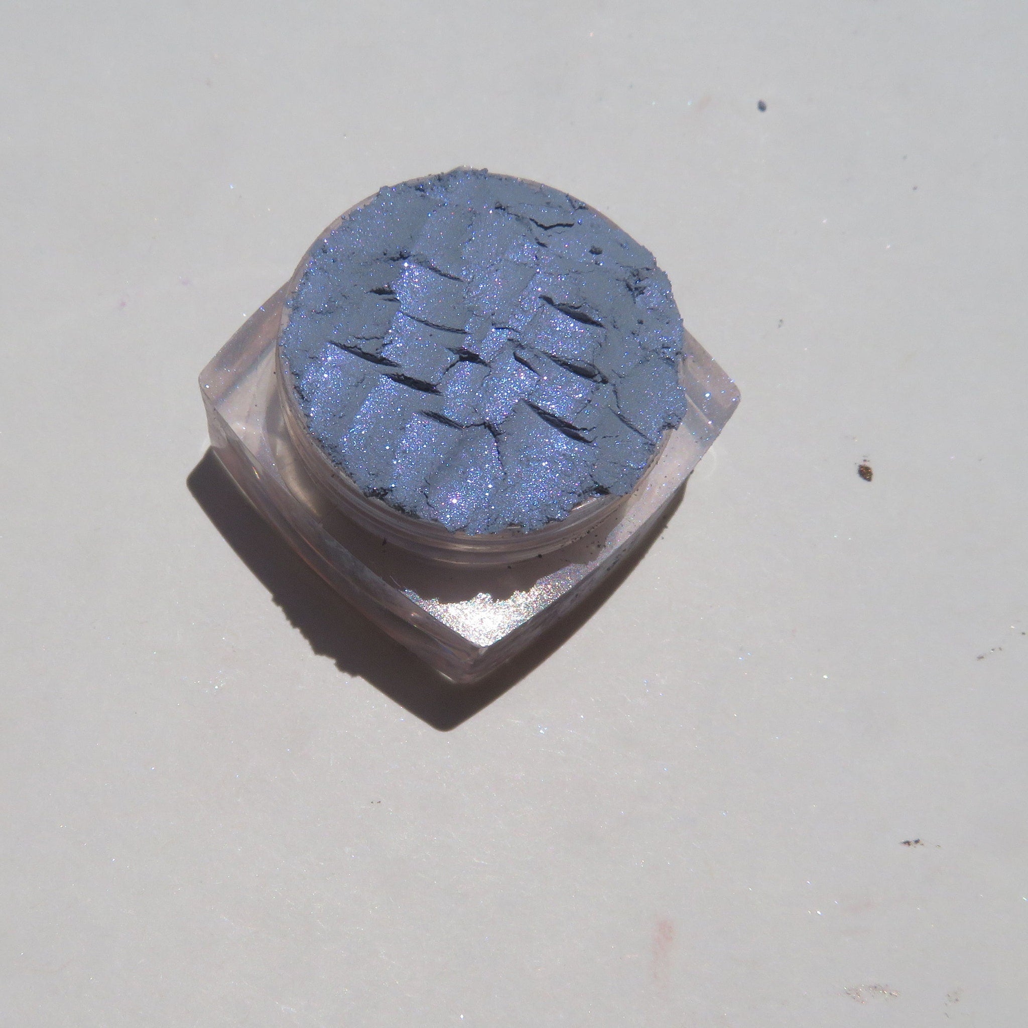 Blue shimmer mineral eyeshadow in clear glass jar with black screw-top lid on marble surface