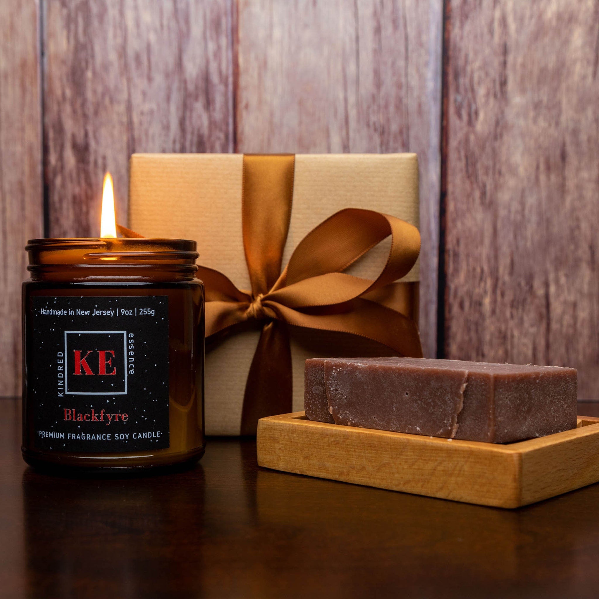 Soy candle with lit flame in amber glass jar, gift box with brown ribbon, and bar soap on wooden tray