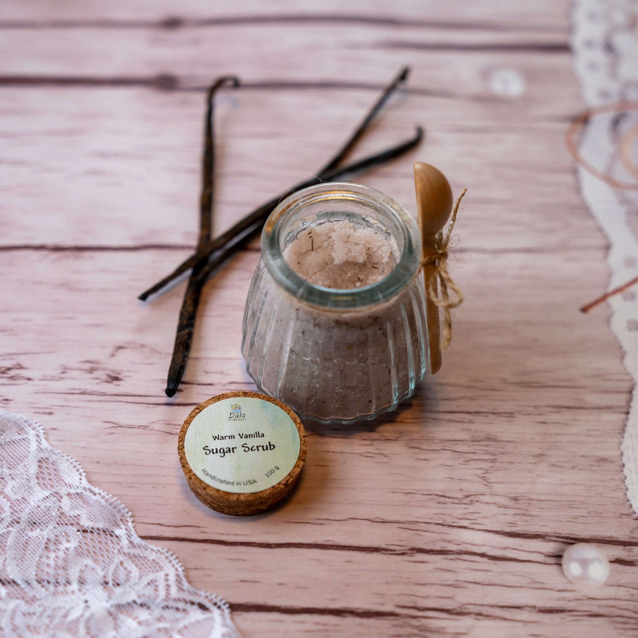 Sugar scrub in glass jar with wooden spoon and vanilla beans on wood surface