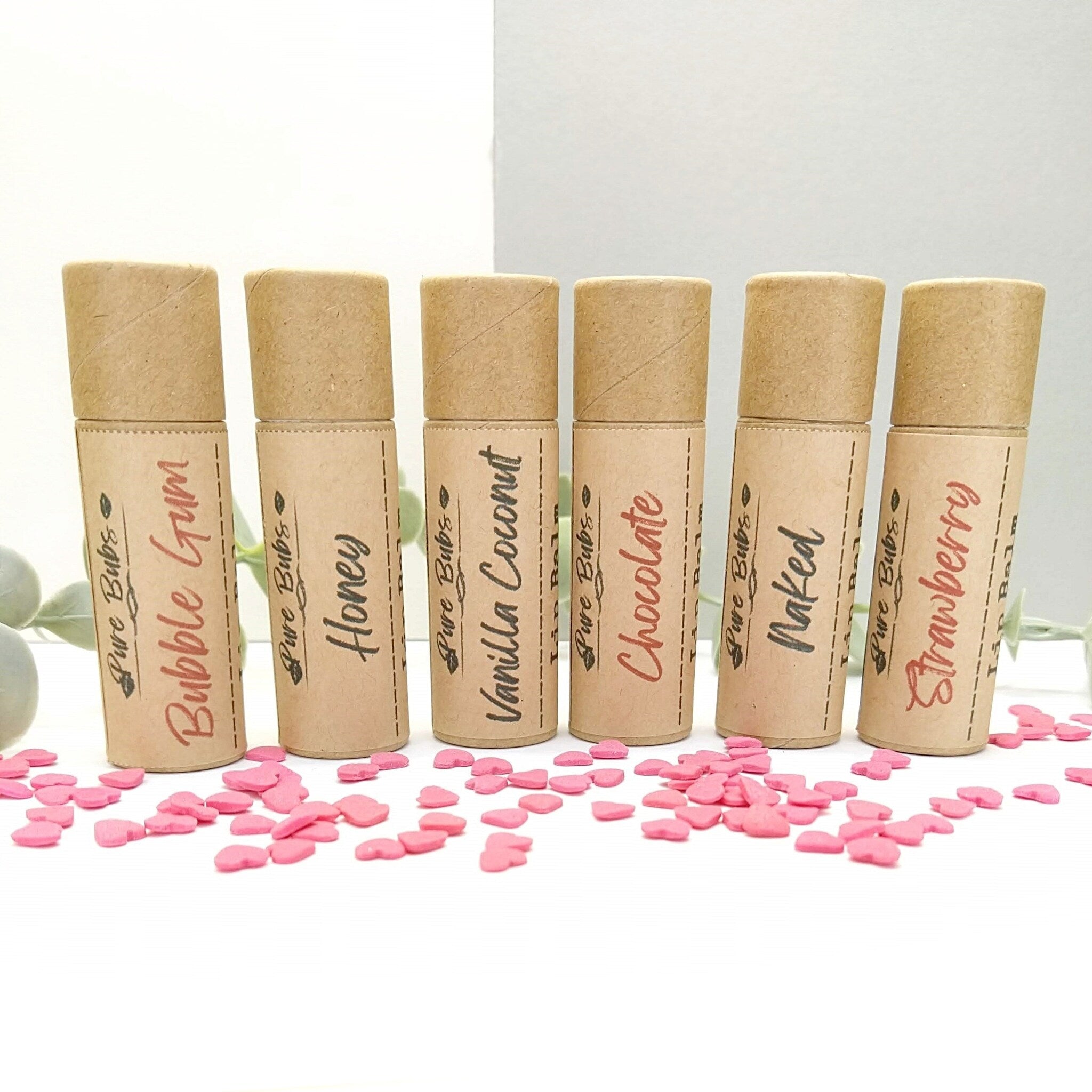 Lip balms in brown paper tubes with flavor labels, pink heart sprinkles around them