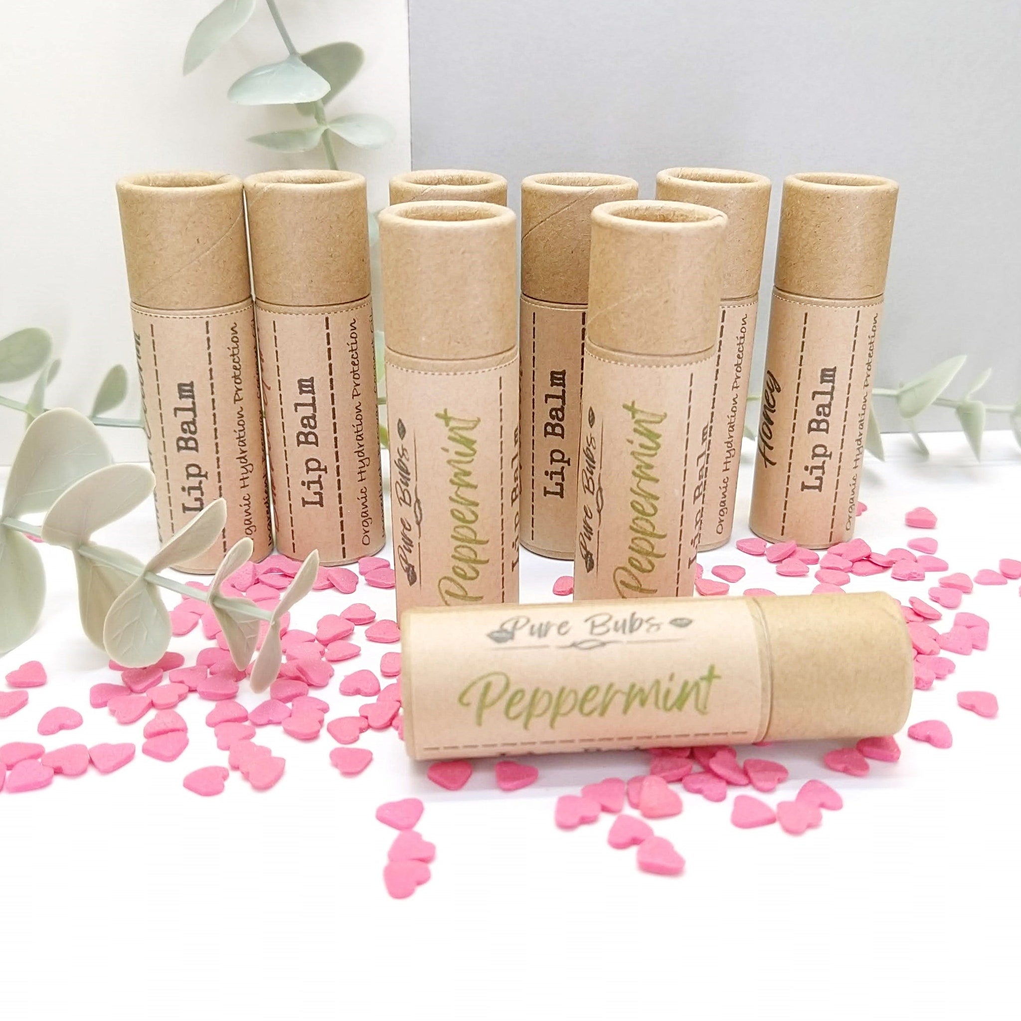 Lip balm tubes, brown cardboard, text label, and pink heart sprinkles on white surface