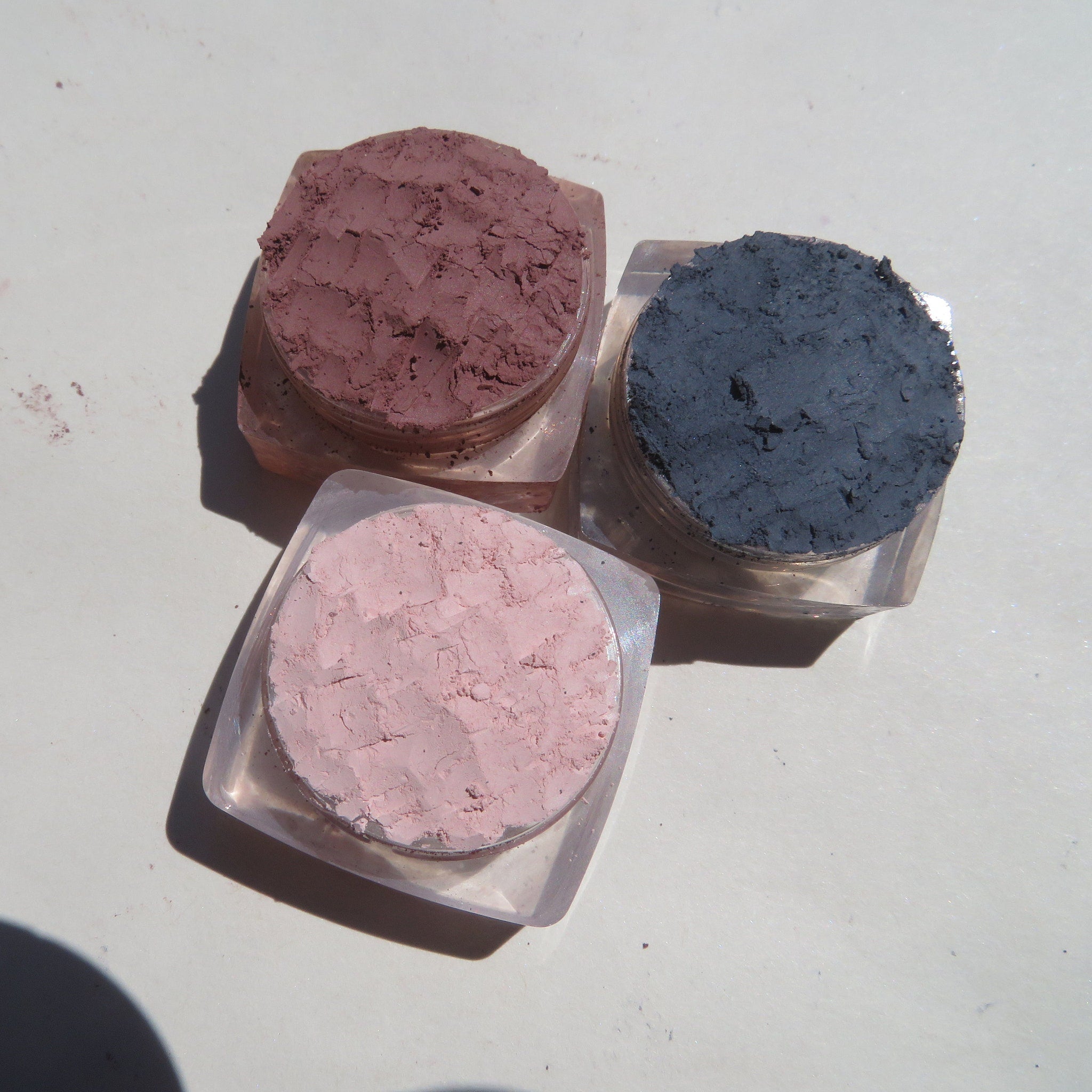 Three matte loose pigment eyeshadows in glass jars with black lids, displayed on a white surface.