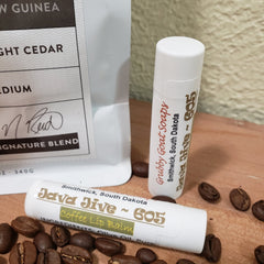 Lip balm tubes with white and gold text, coffee beans on wood surface, Grubby Goat Soaps brand