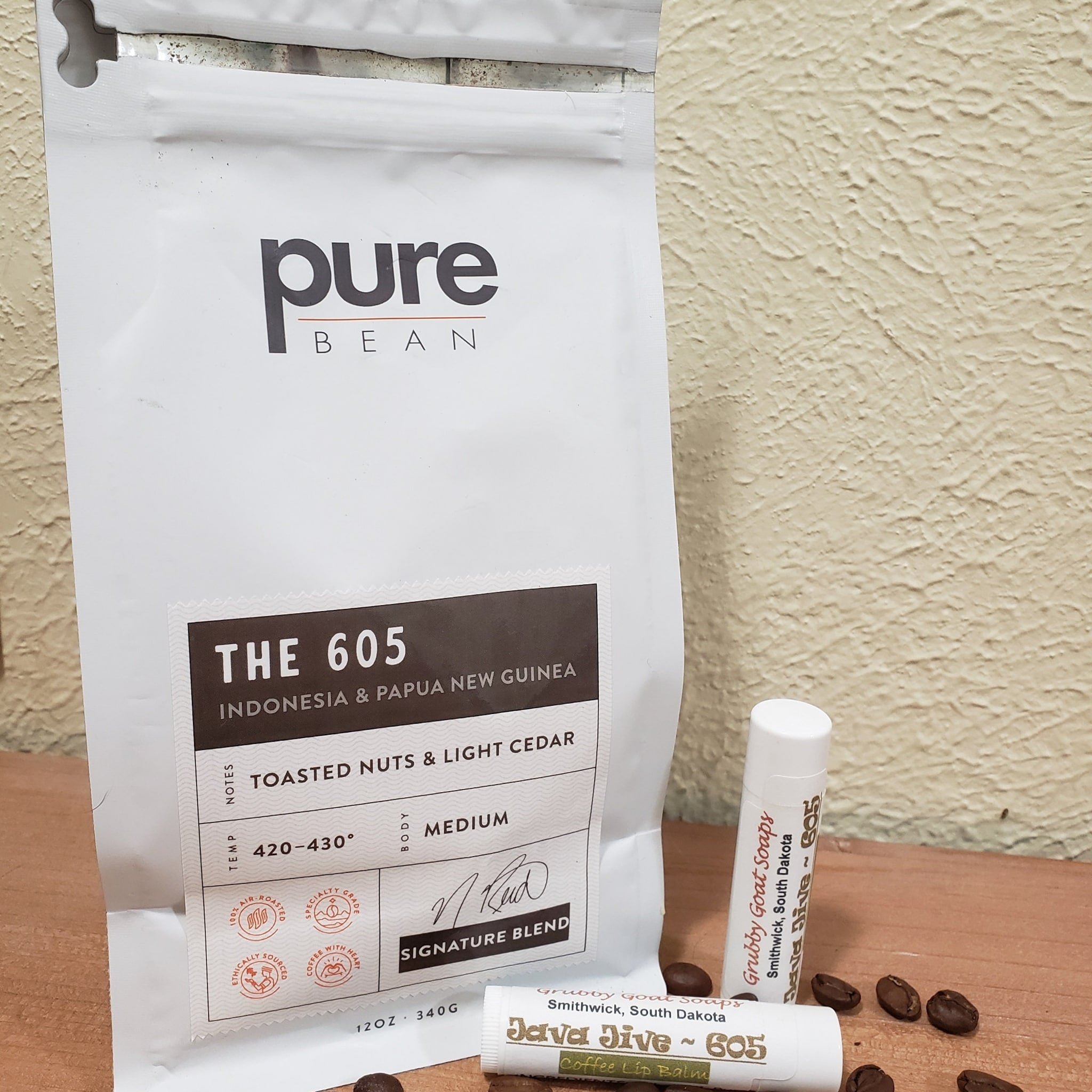 Coffee beans and lip balm on wood surface, white bag with "pure bean" and "THE 605" text visible