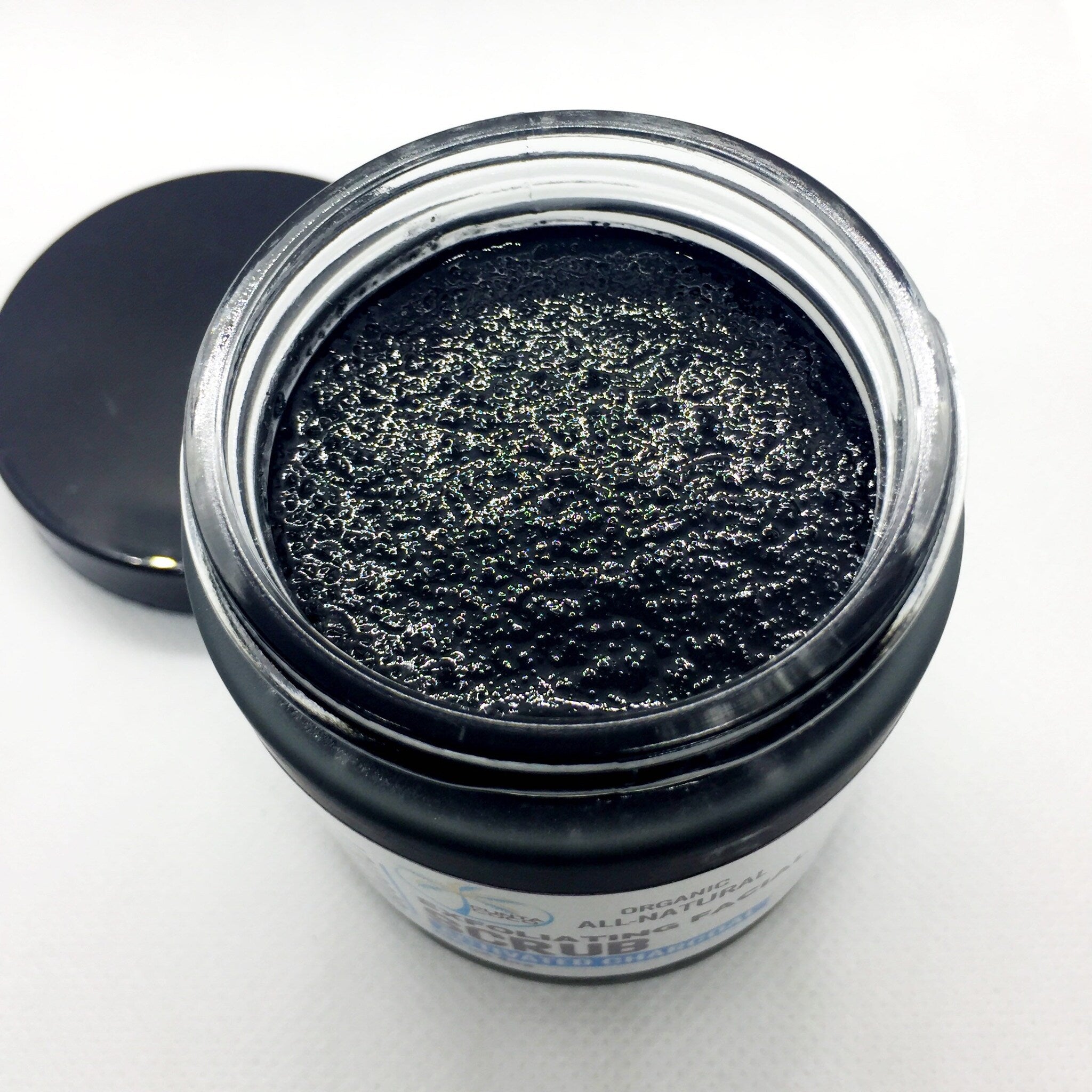 Exfoliating face scrub in a clear glass jar with black lid, dark granular texture, and brand label visible