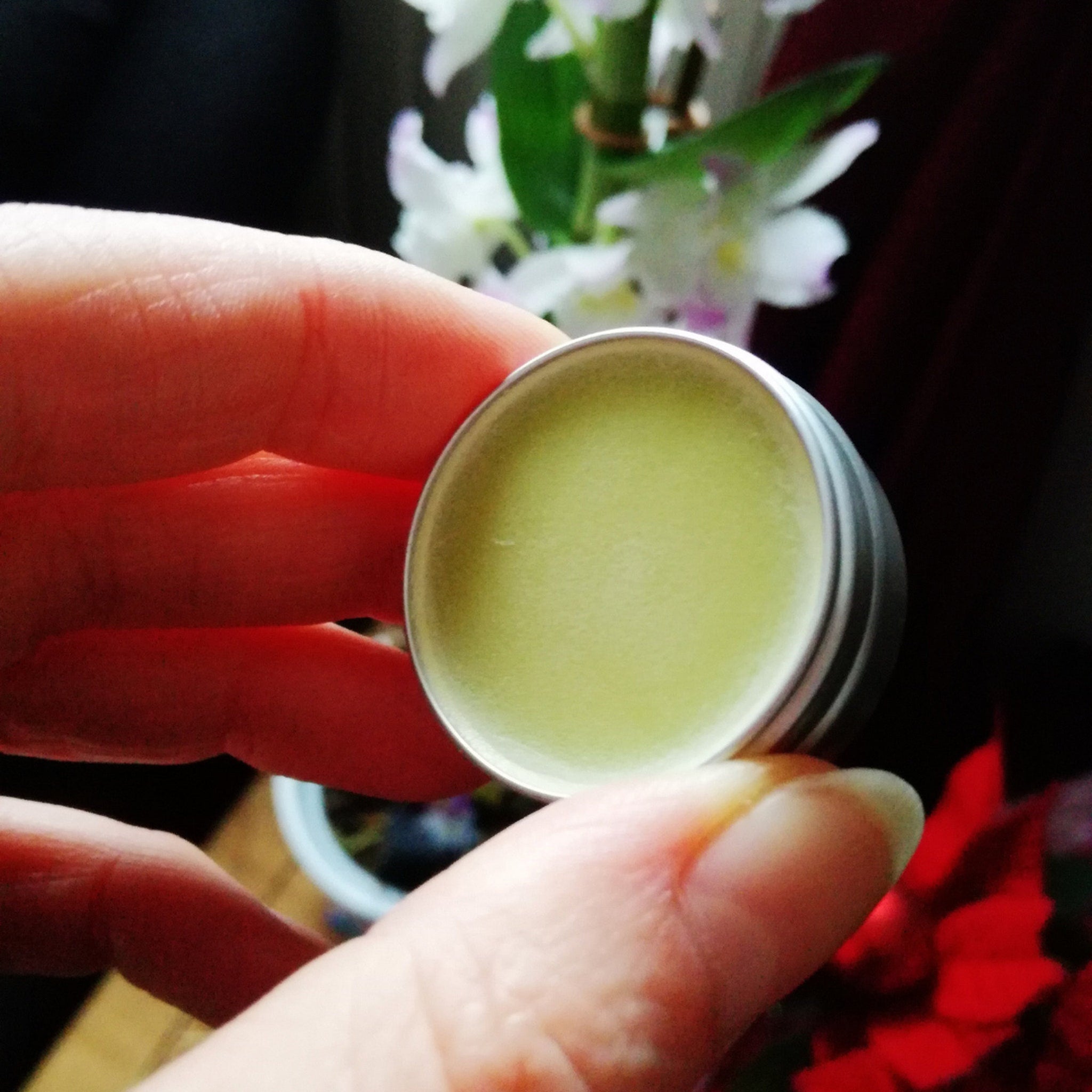 Lip balm in silver tin with light green balm, held by hand, with flowers in background
