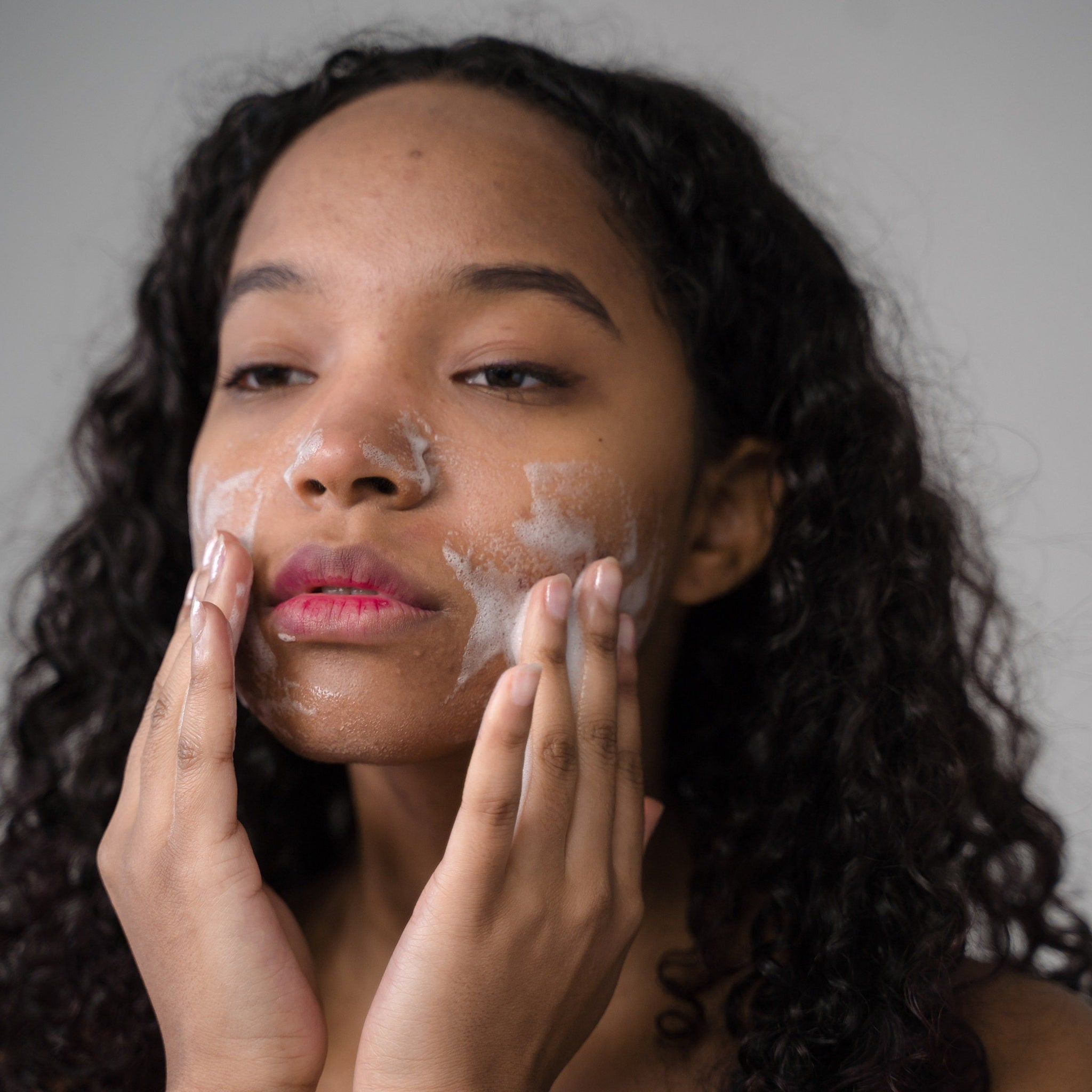 Facial cleanser application, woman with dark curly hair and white foam on face, hands touching skin