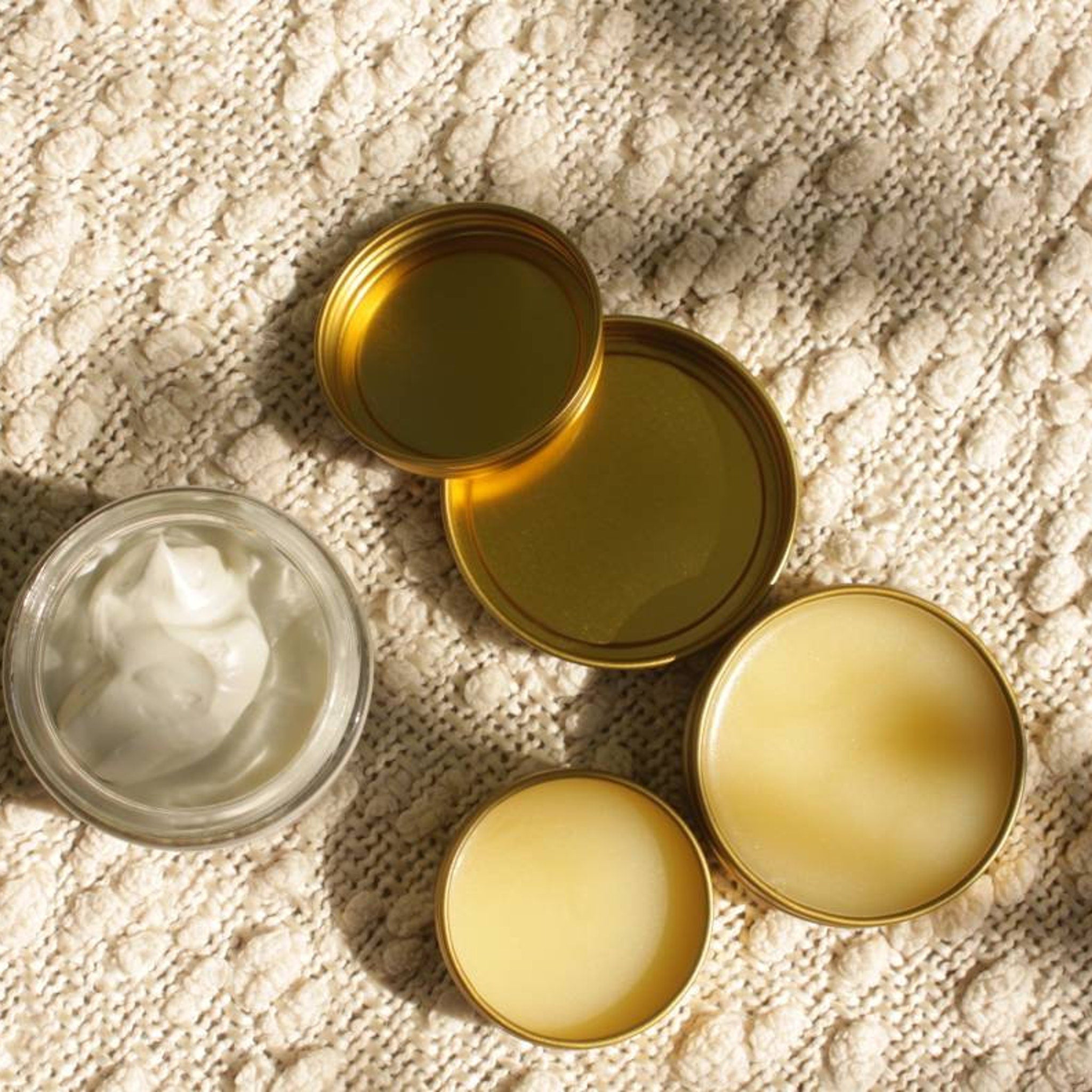 Skincare products with gold lids, white cream, and yellow balm on textured beige fabric