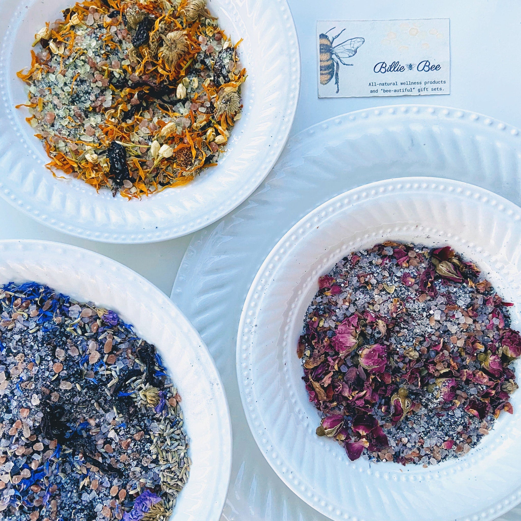 Bath salts with orange, blue, and pink botanicals in white bowls, Billie Bee products