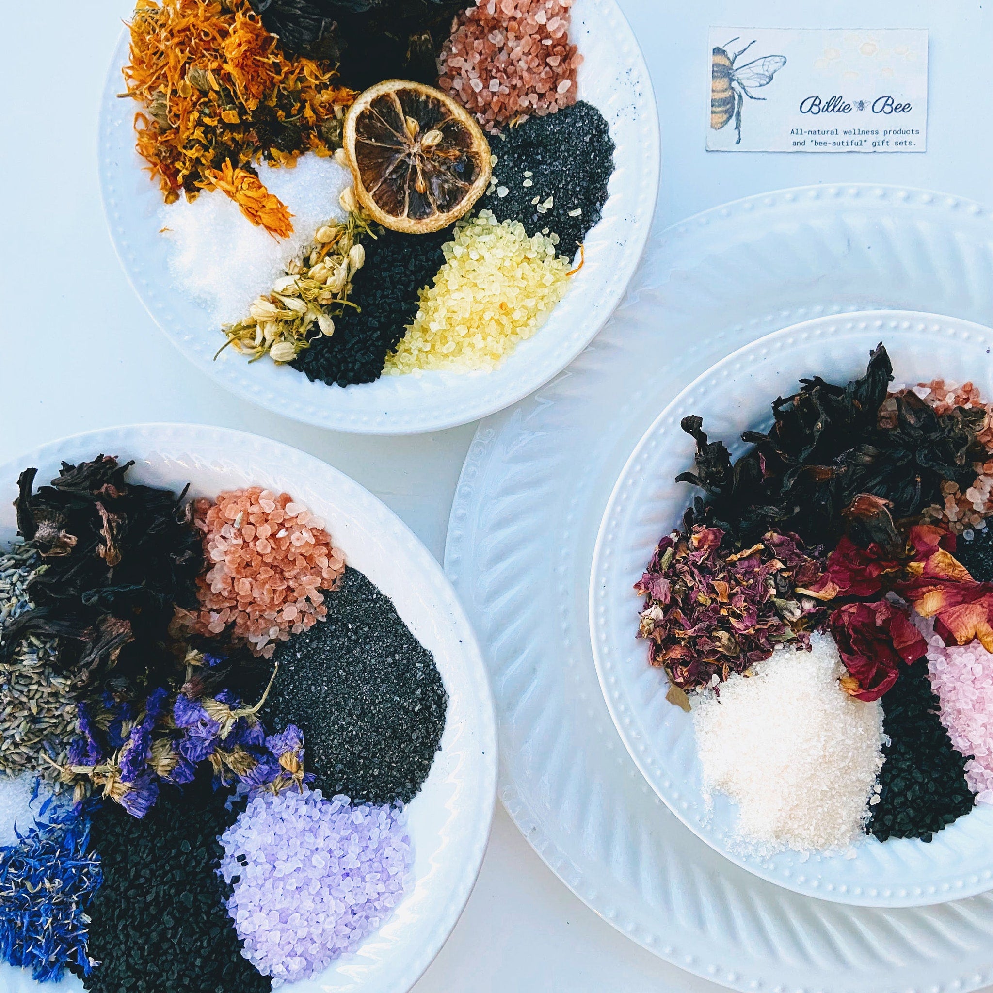 Bath salts on white plates with dried flowers, salt crystals, and Billie Bee branding