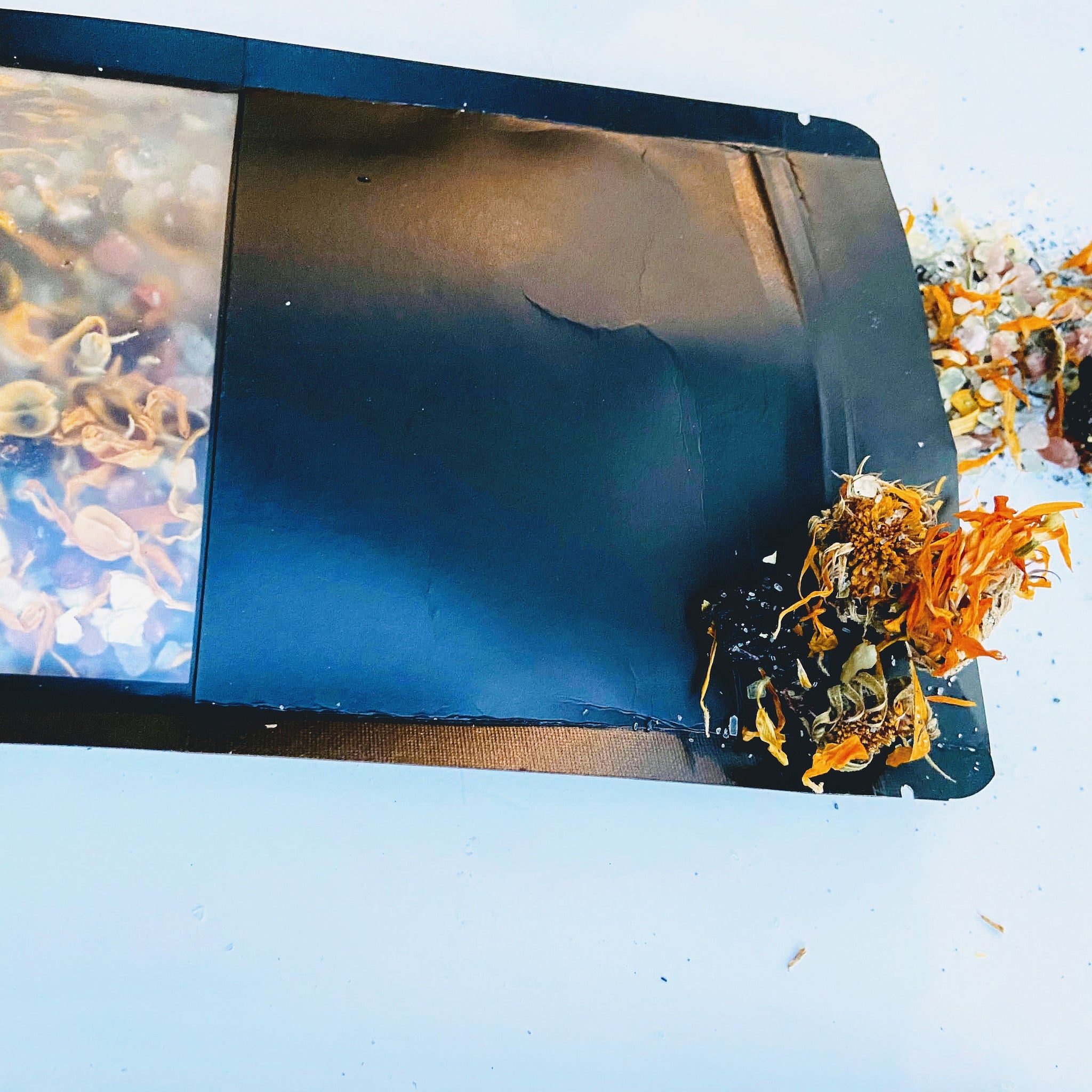Black packet with clear window showing dried herbs, orange and yellow plant matter, on white surface