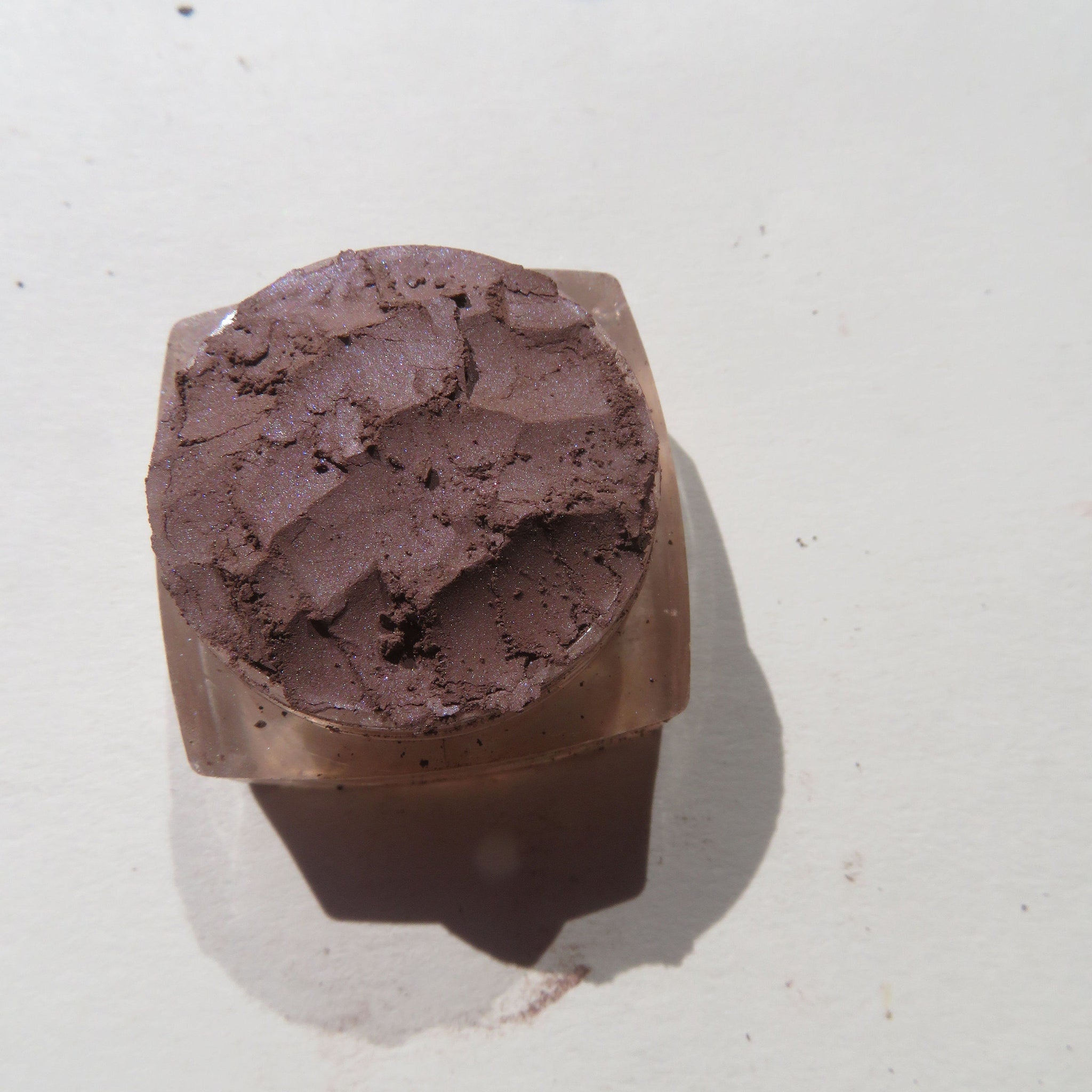 Brown eyeshadow in a clear plastic container with a textured surface and shadow