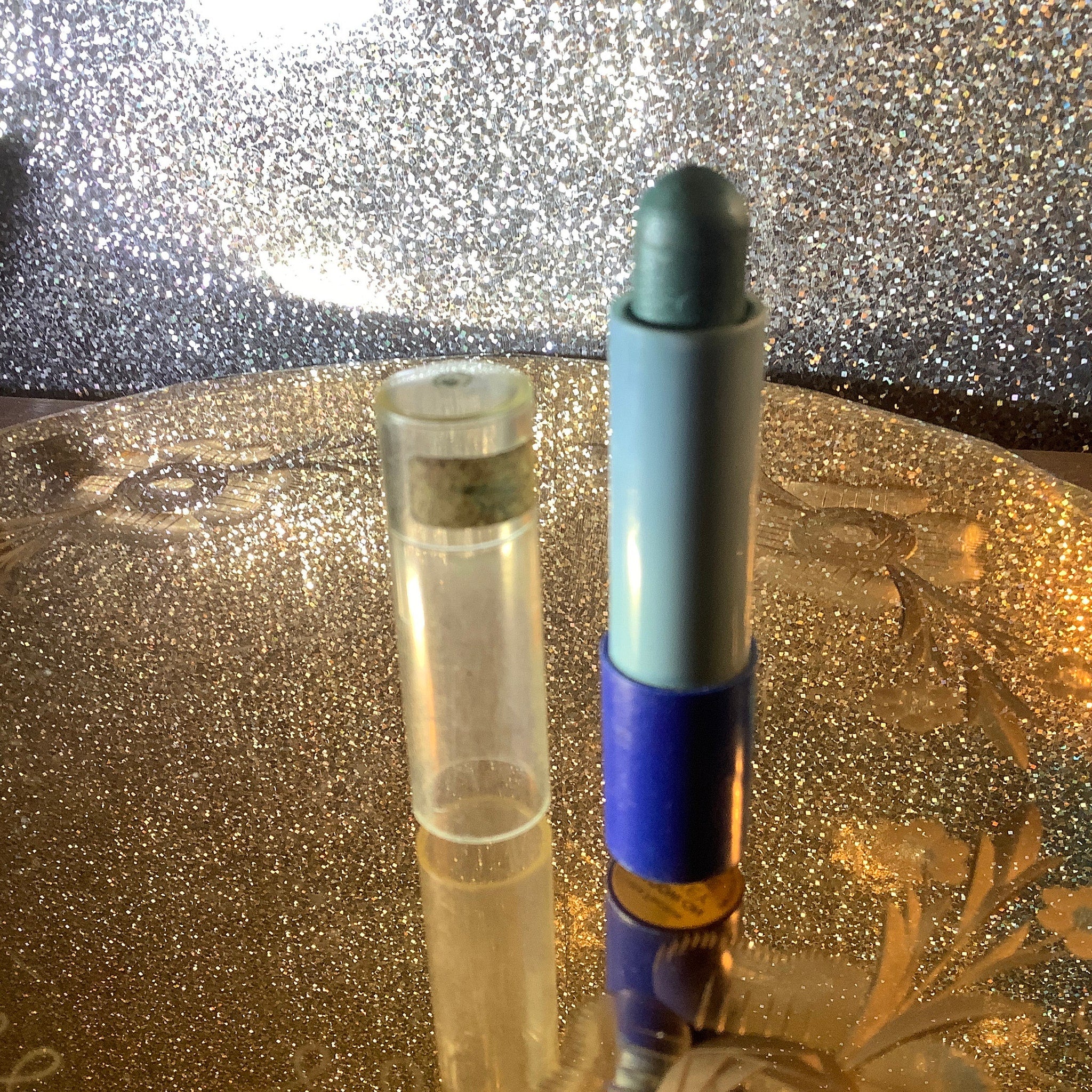 Lipstick tube with blue, green, and clear plastic, sitting on gold glitter surface.