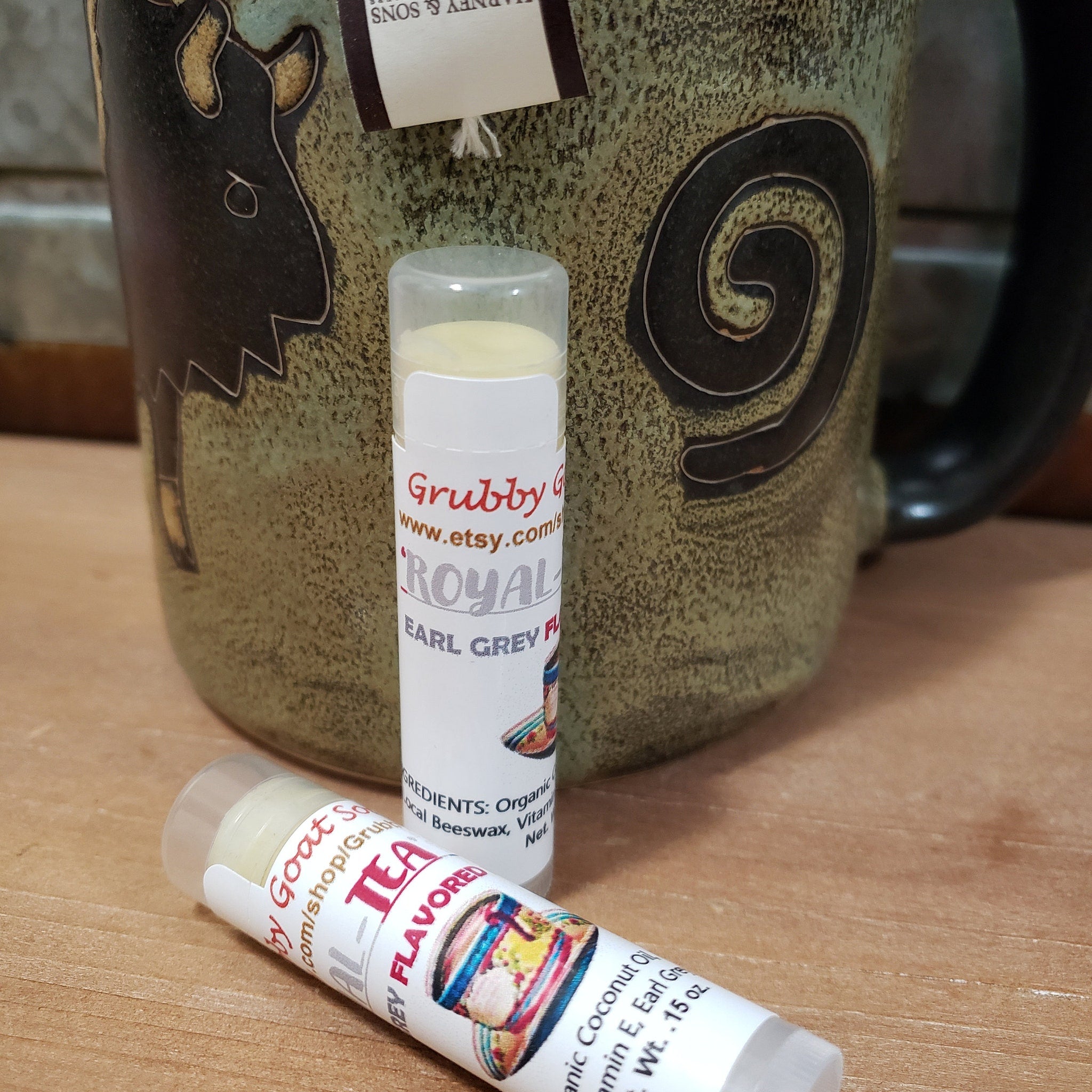 Lip balm tubes with white labels, clear caps, and "Grubby Goat" text, next to a mug