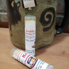 Lip balm tubes with white labels, clear caps, and "Grubby Goat" text, next to a mug