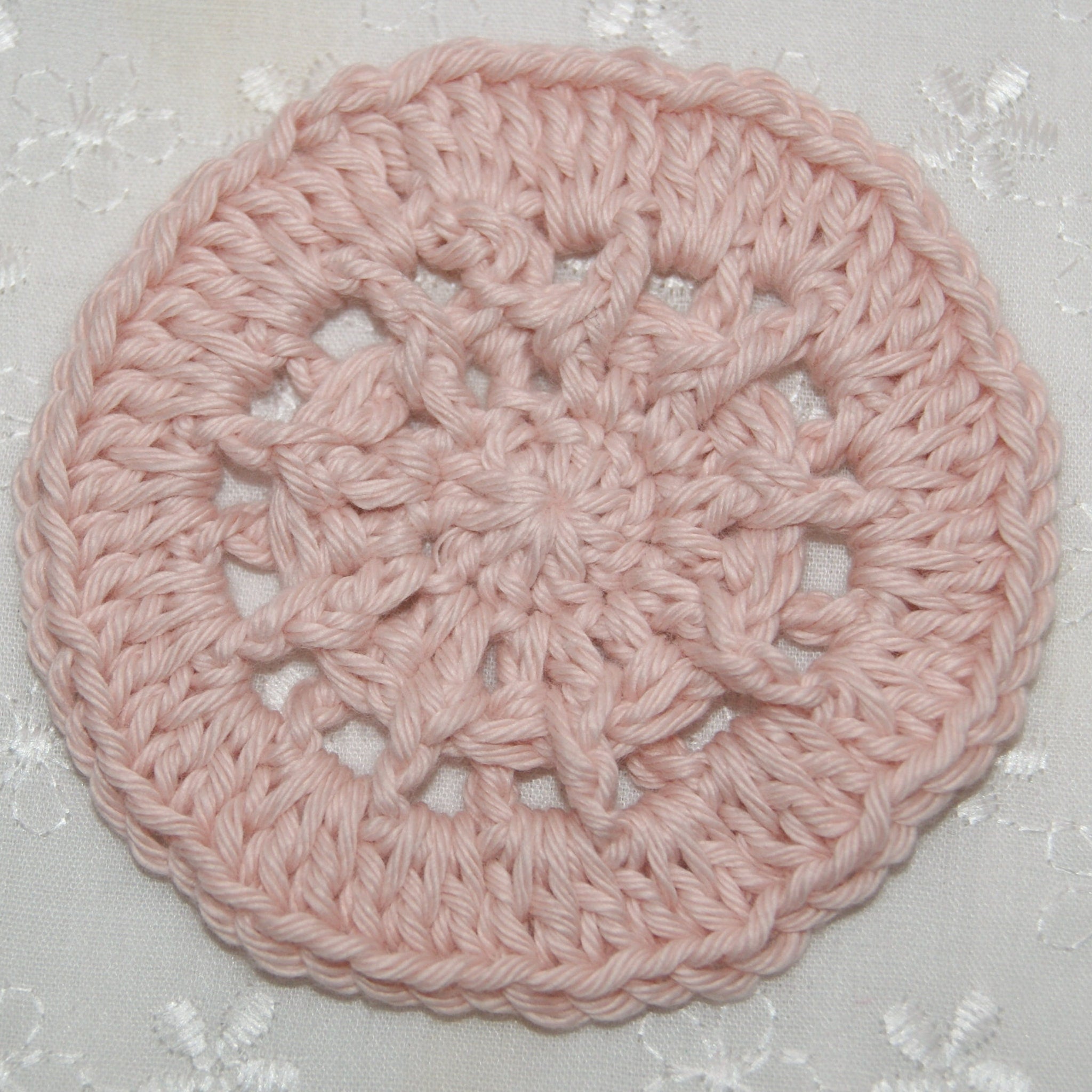 Crochet coaster in pale pink cotton with openwork pattern on white fabric