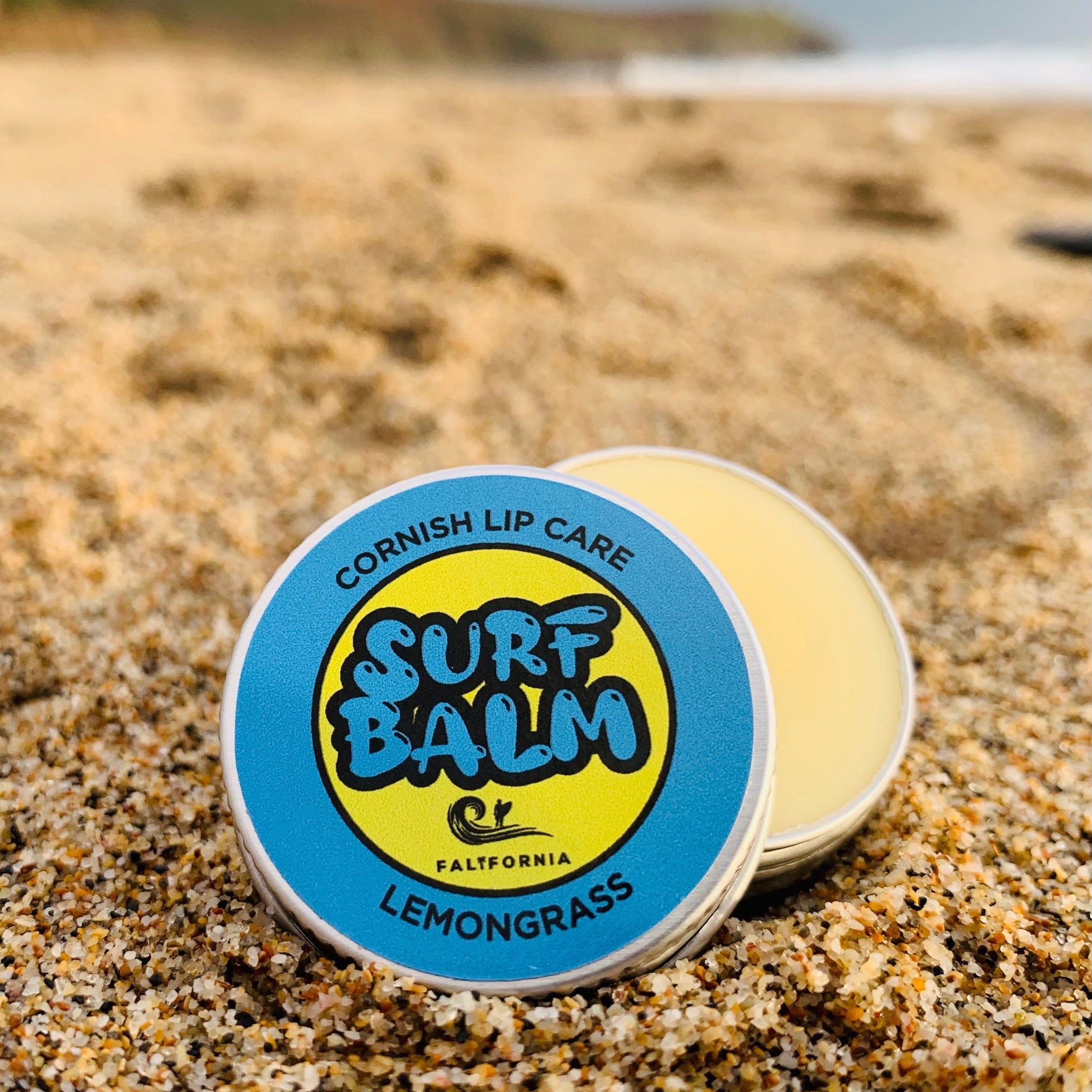 Lip balm tin with blue, yellow, and white label on sand, "Surf Balm" text visible