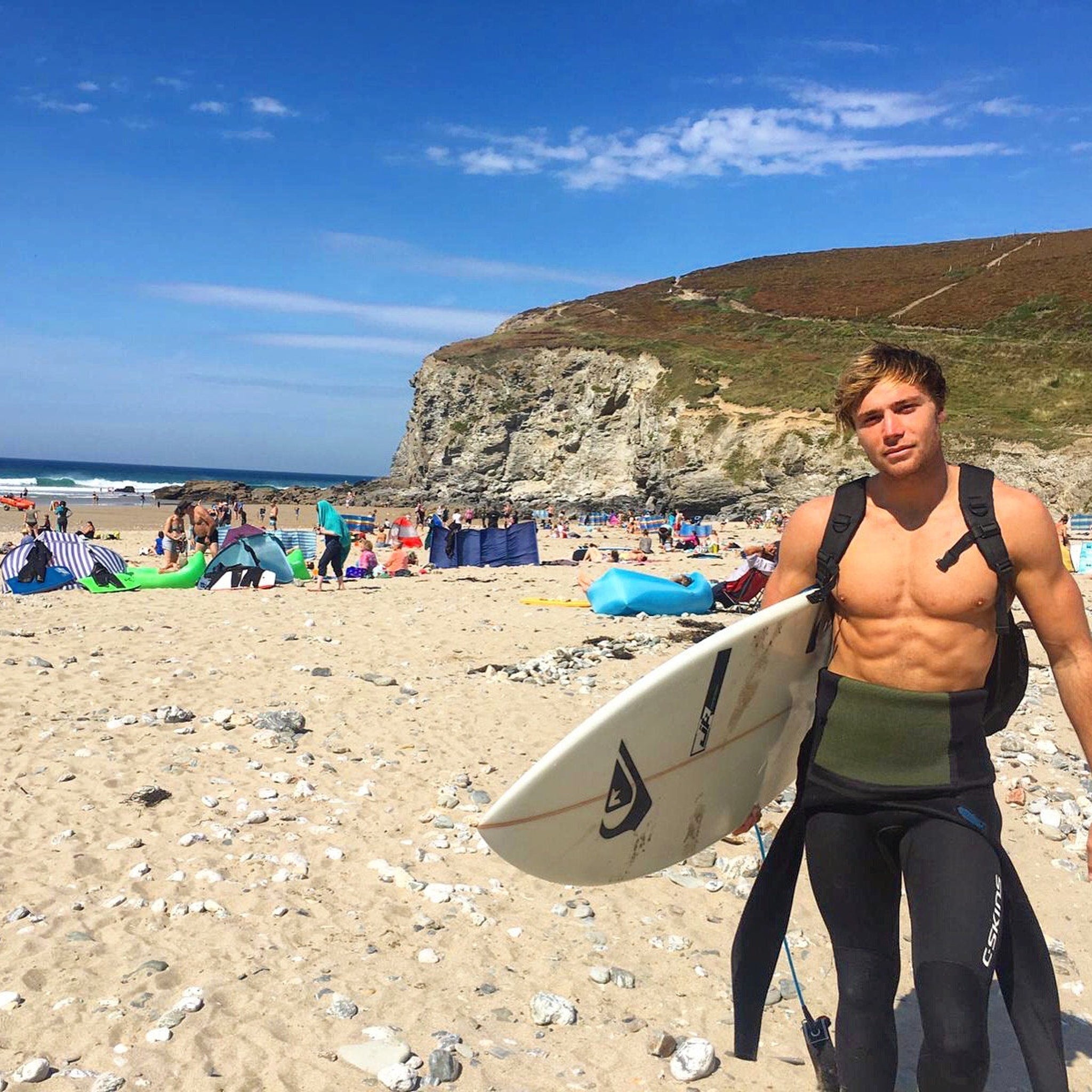 Surfer holding surfboard, wearing black and green wetsuit, on beach
