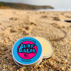 Lip balm tin with blue and pink label, open on sand, "Surf Balm" text visible