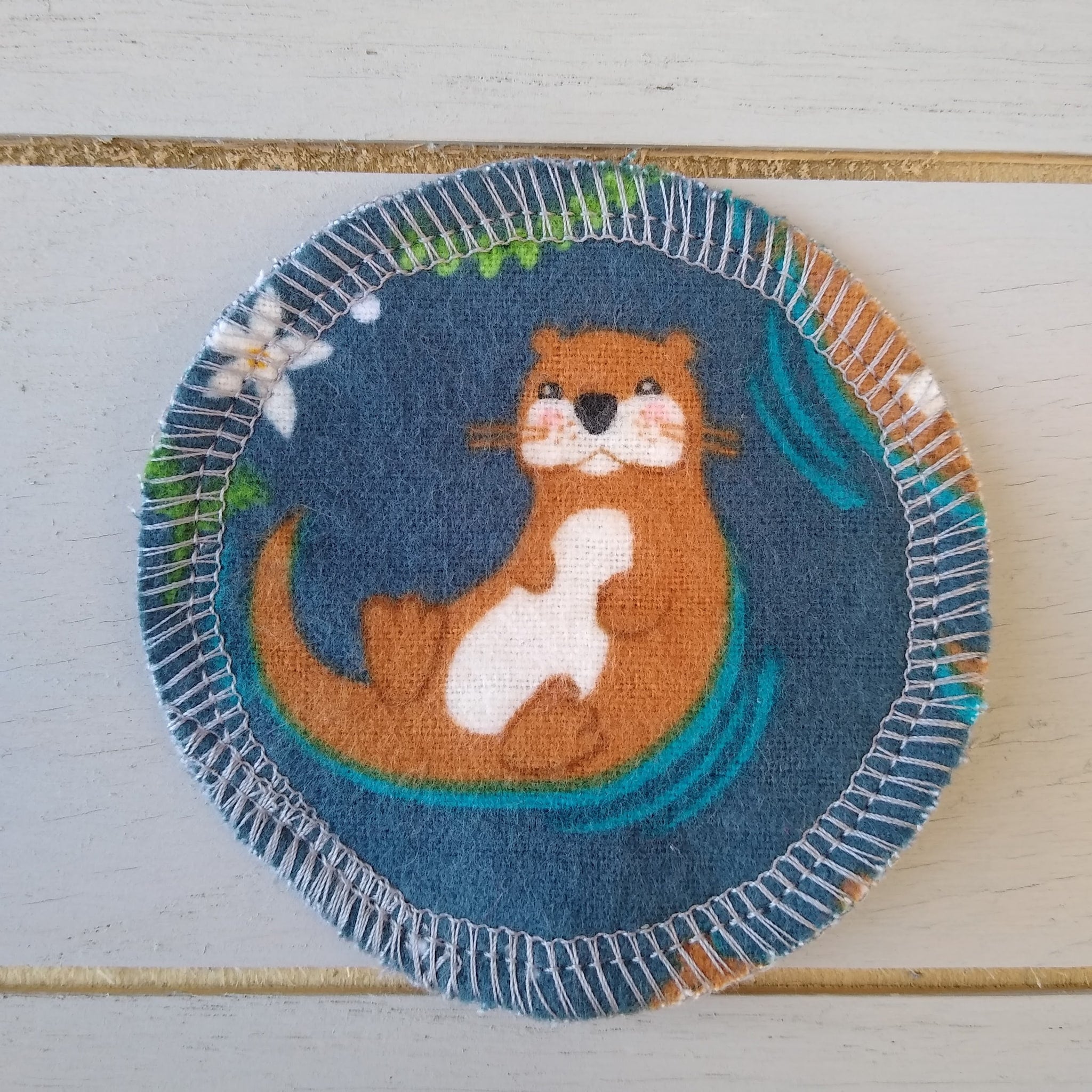 Coaster with otter print, blue fabric, white stitched edge, and floral accents