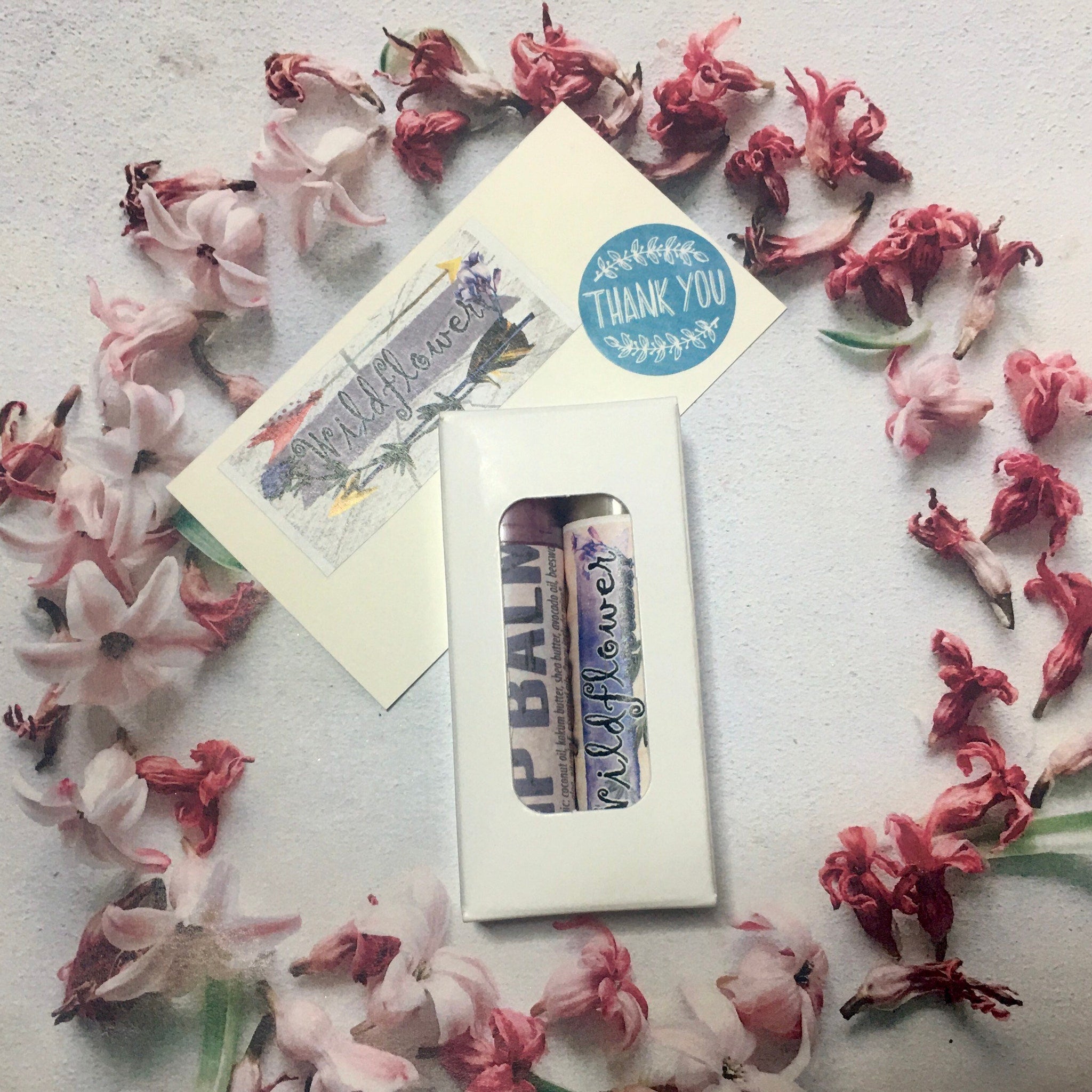 Lip balm tubes with wildflower print, white box, and floral arrangement on white surface