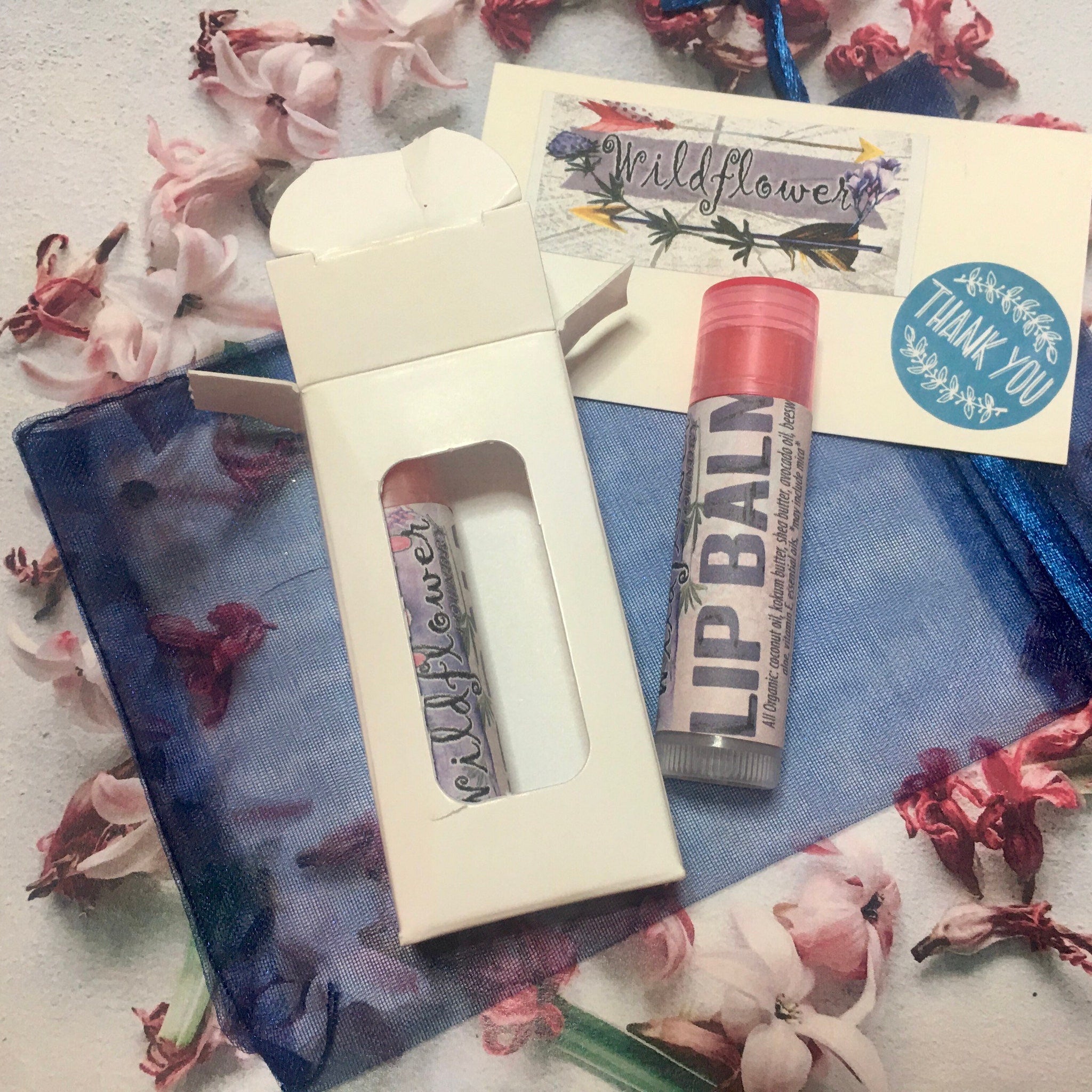 Lip balm tube with pink cap and label, in white box with wildflower text, on blue mesh fabric