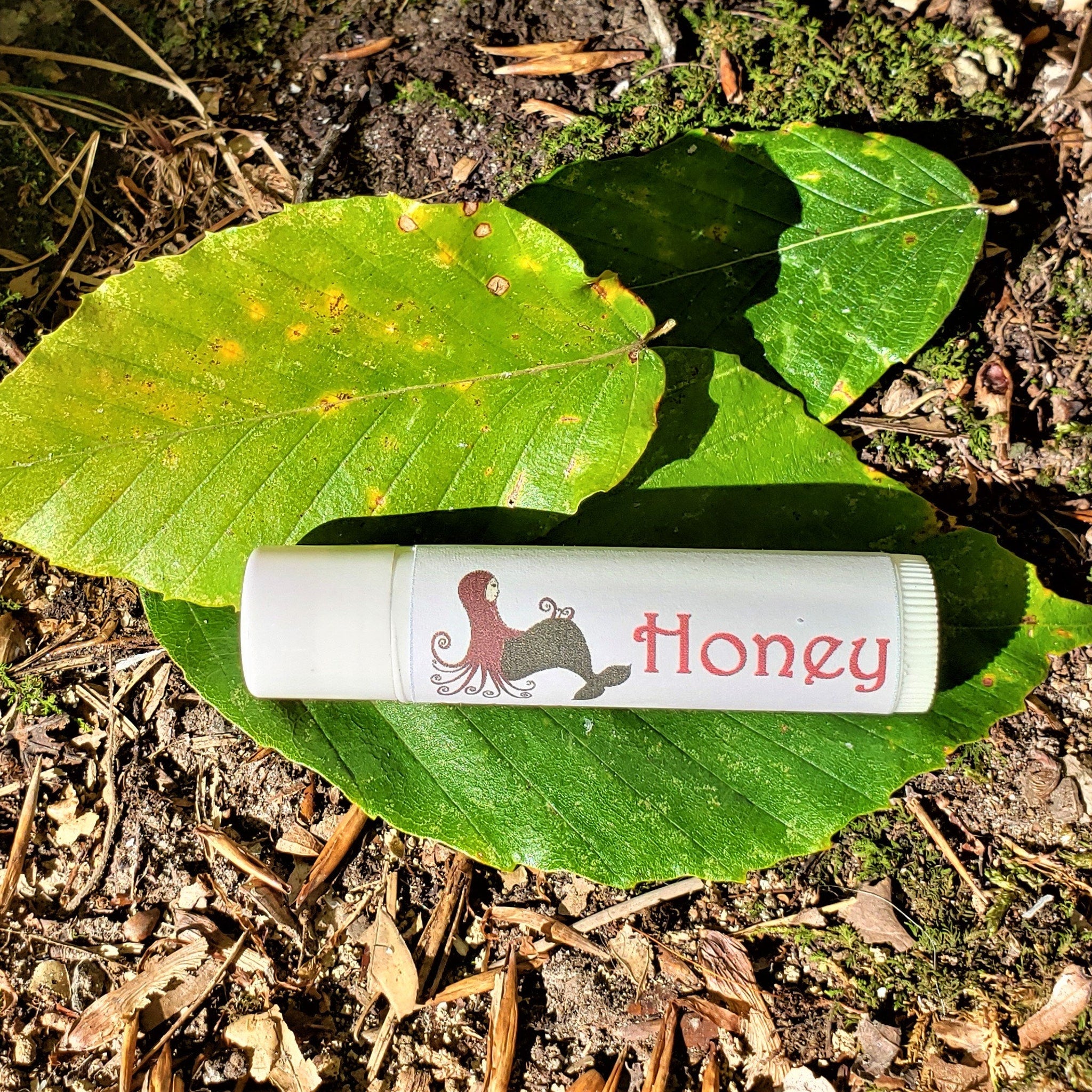Lip balm tube with white and red text, on green leaf, with Honey text visible