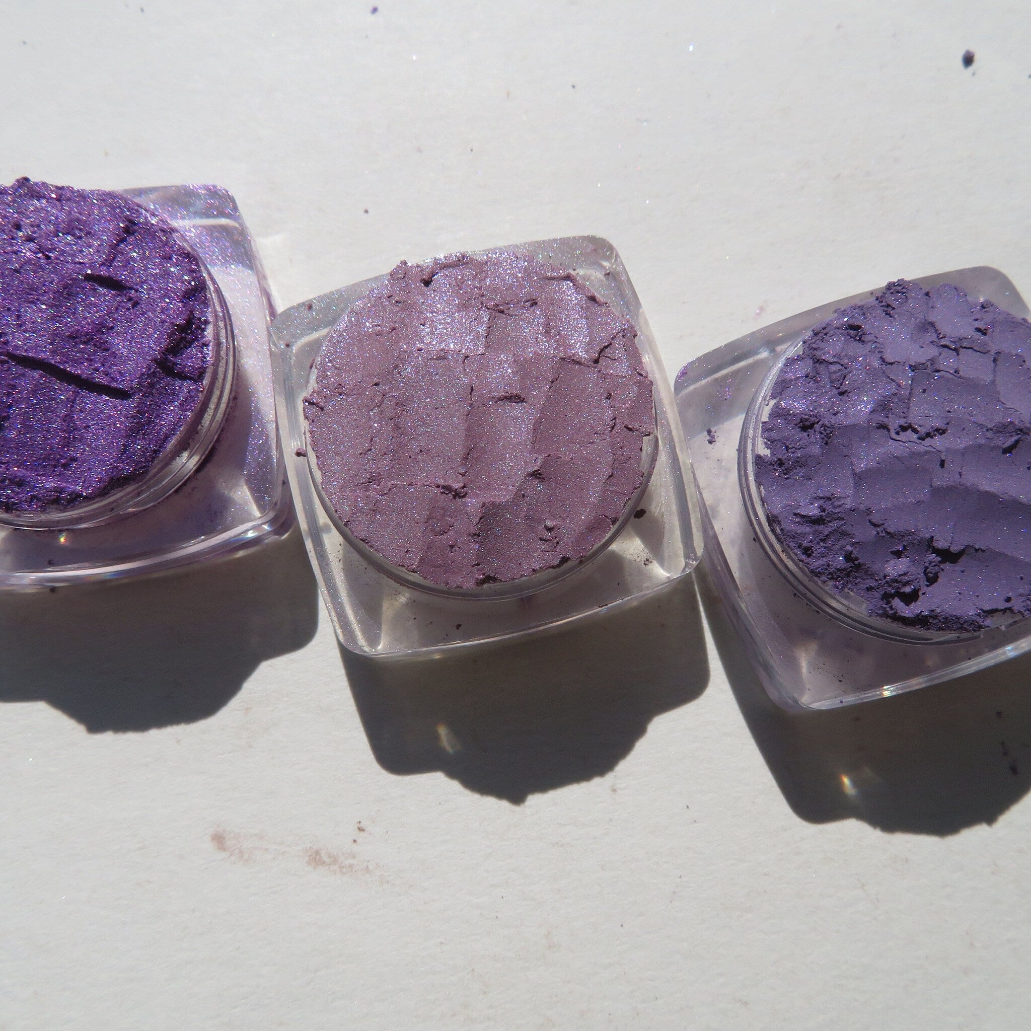 Three loose purple mineral eyeshadows shimmering in glass jars with gold lids, displayed on marble surface.