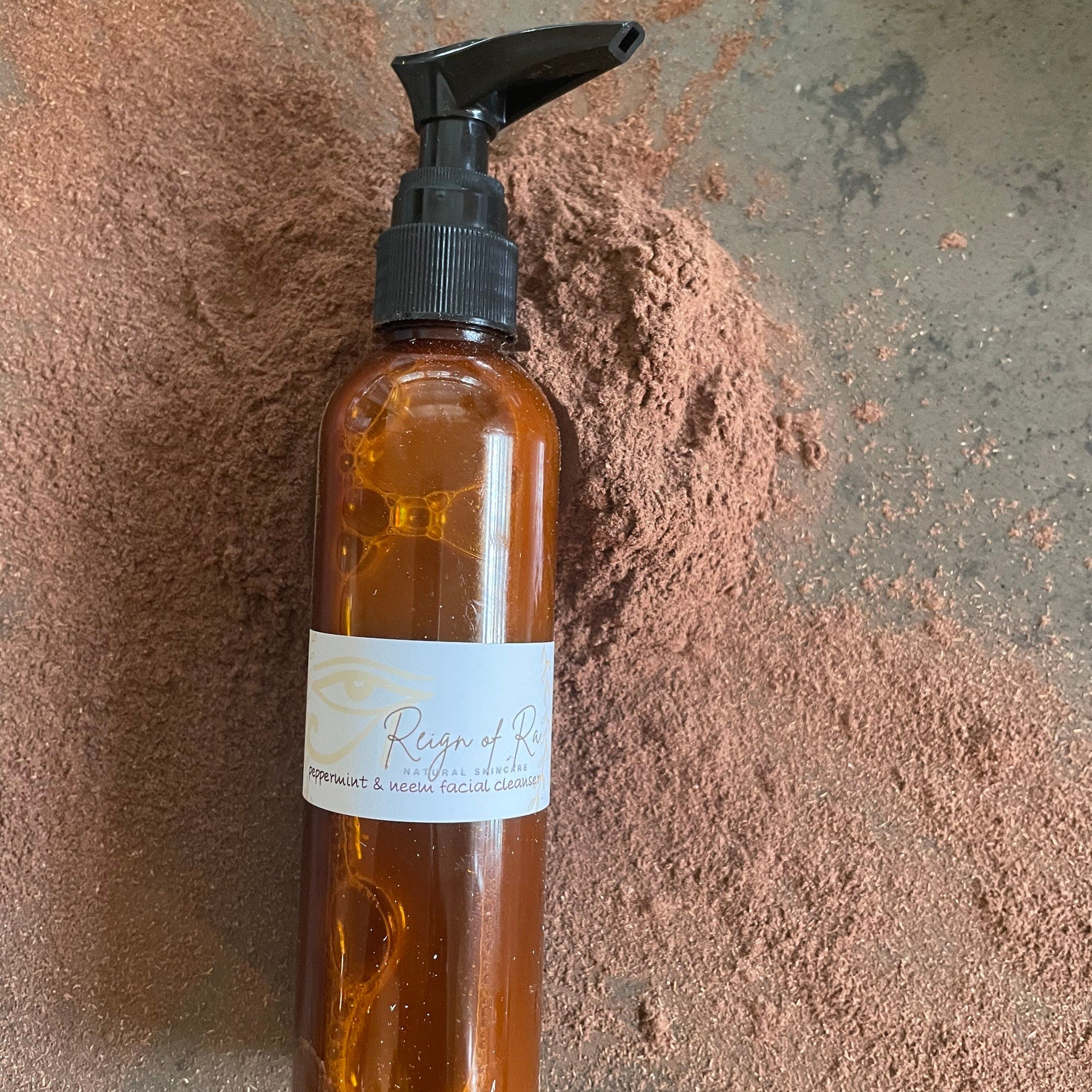 Facial cleanser bottle, brown glass, black pump, Reign of Ra label, on brown powder