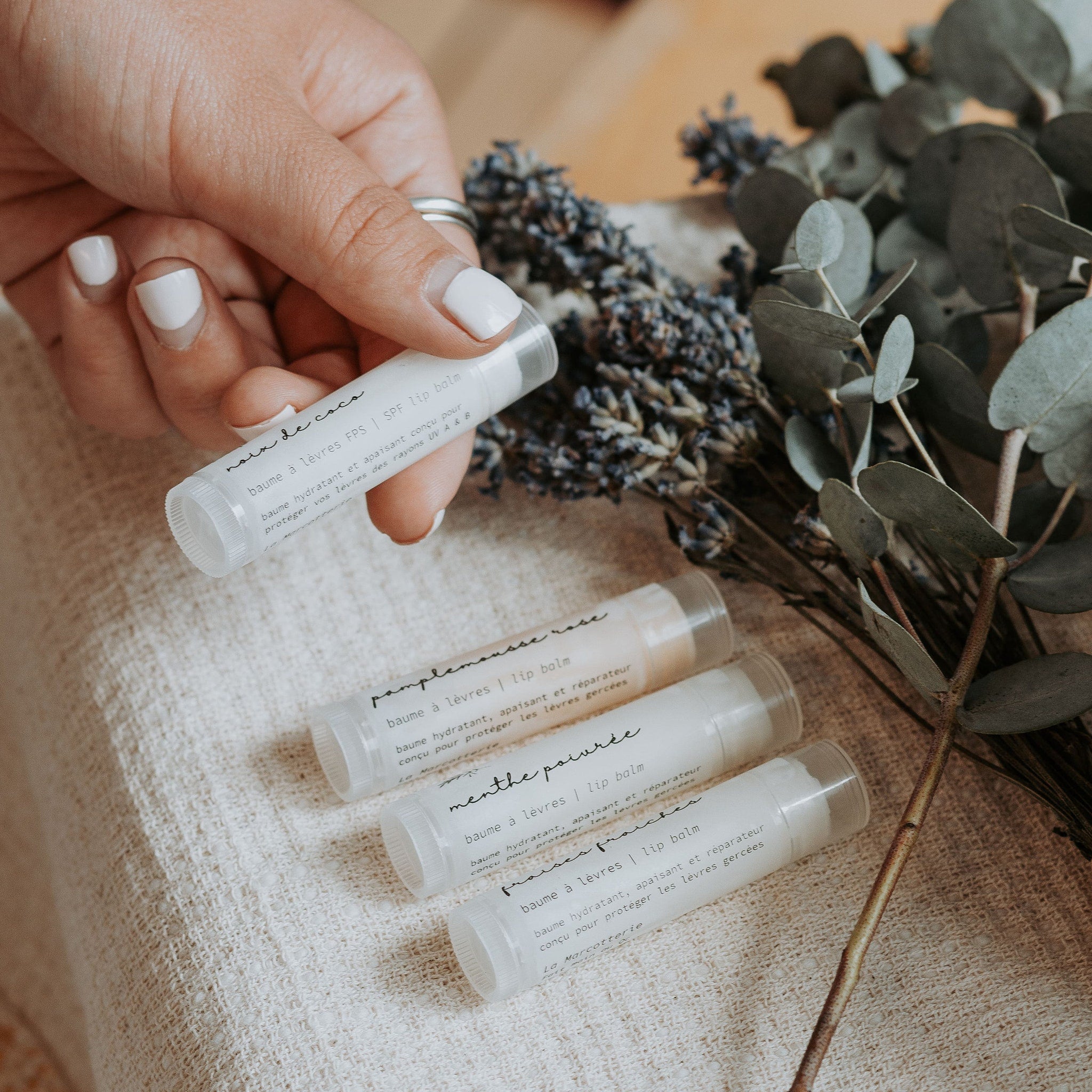 Lip balm tubes with white labels, clear caps, and text, held by hand with white nails