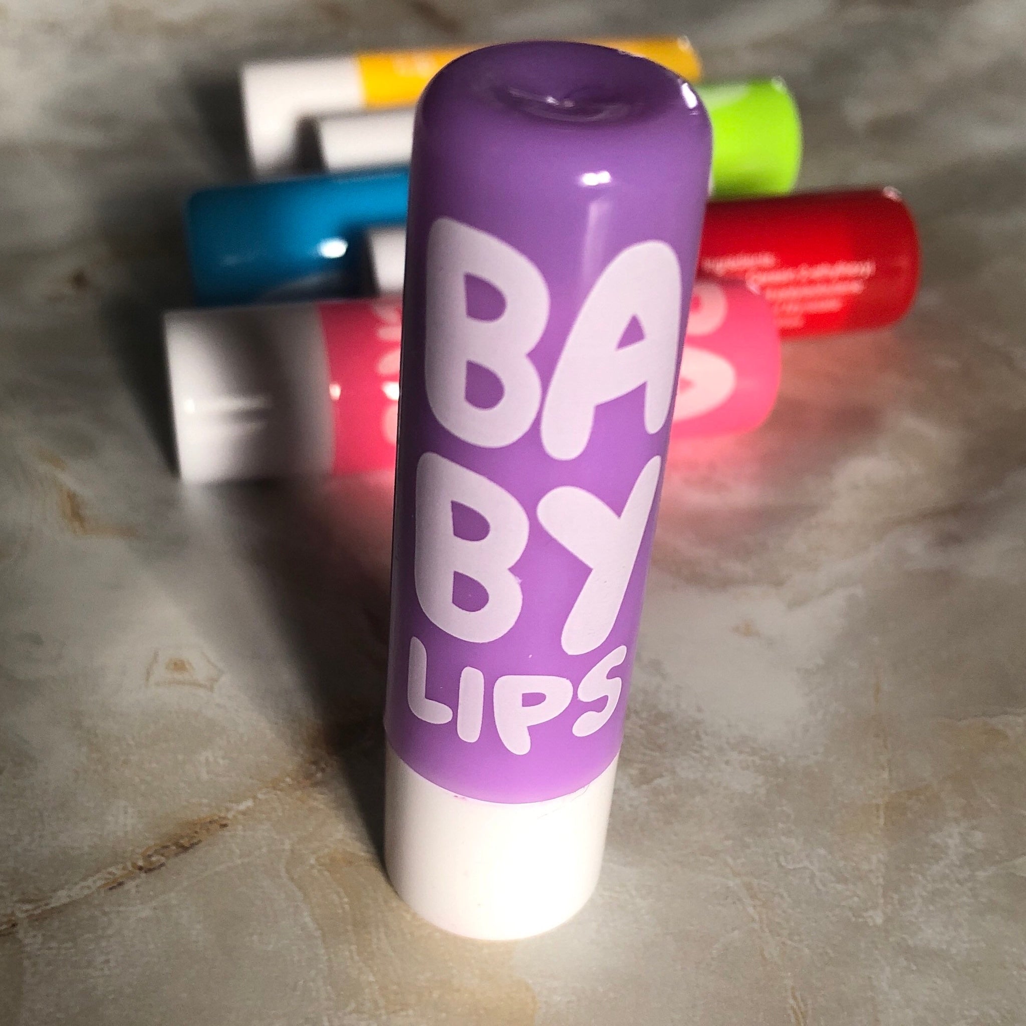 Lip balm tube, purple and white with "BABY LIPS" text, other colorful tubes in background