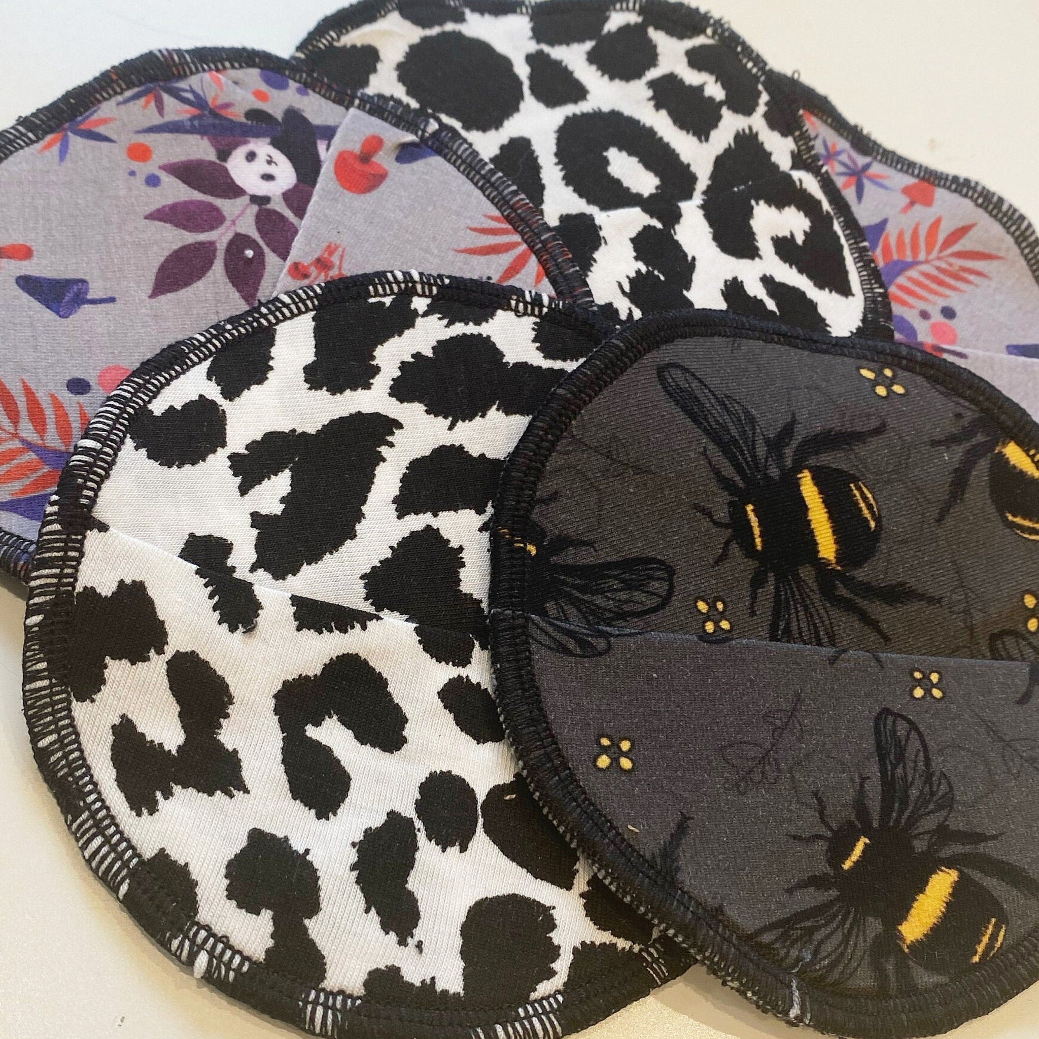 Reusable nursing pads with various patterns, including black and white spots, bees, and floral designs, with black trim