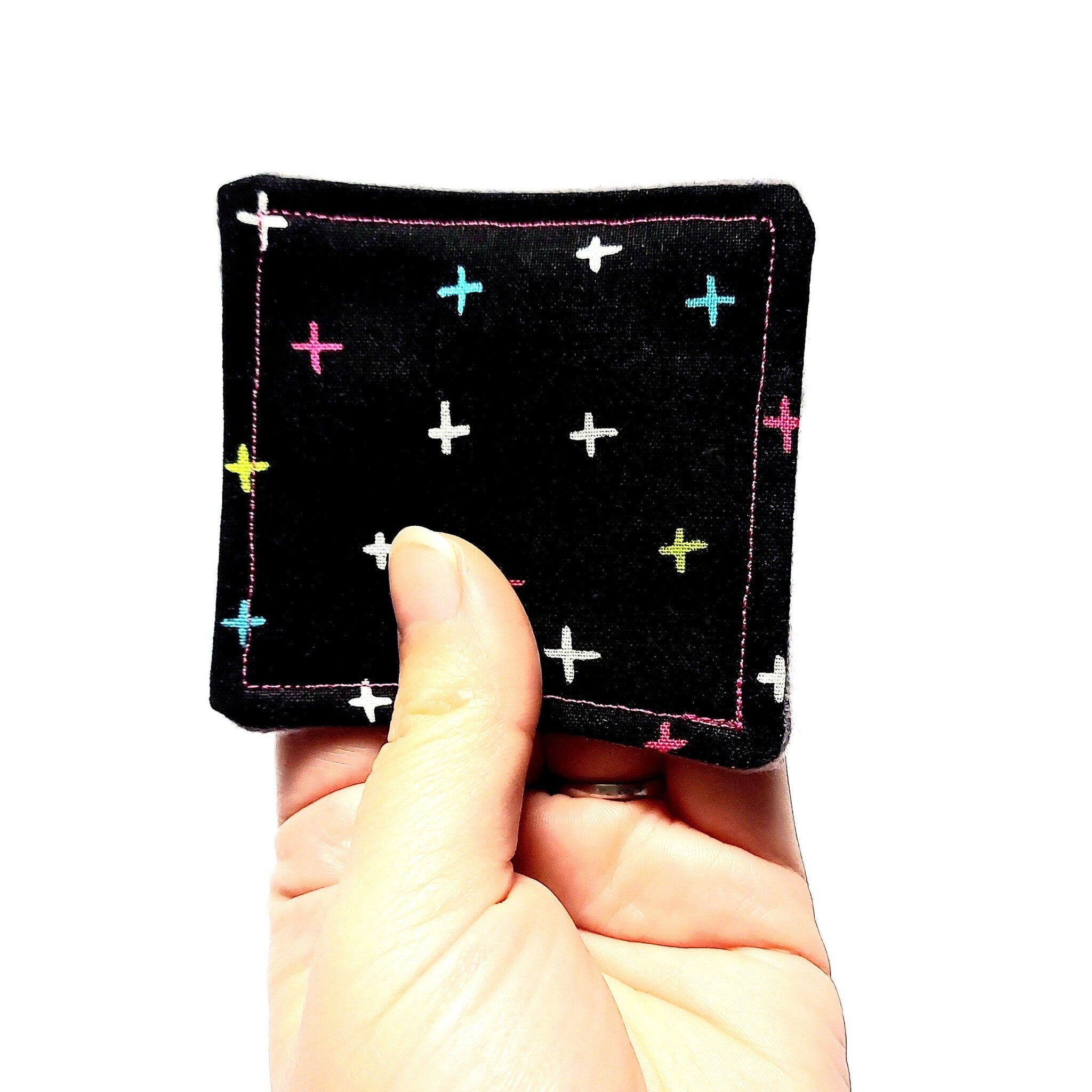 Black square fabric with colorful cross pattern held in hand, pink stitching detail