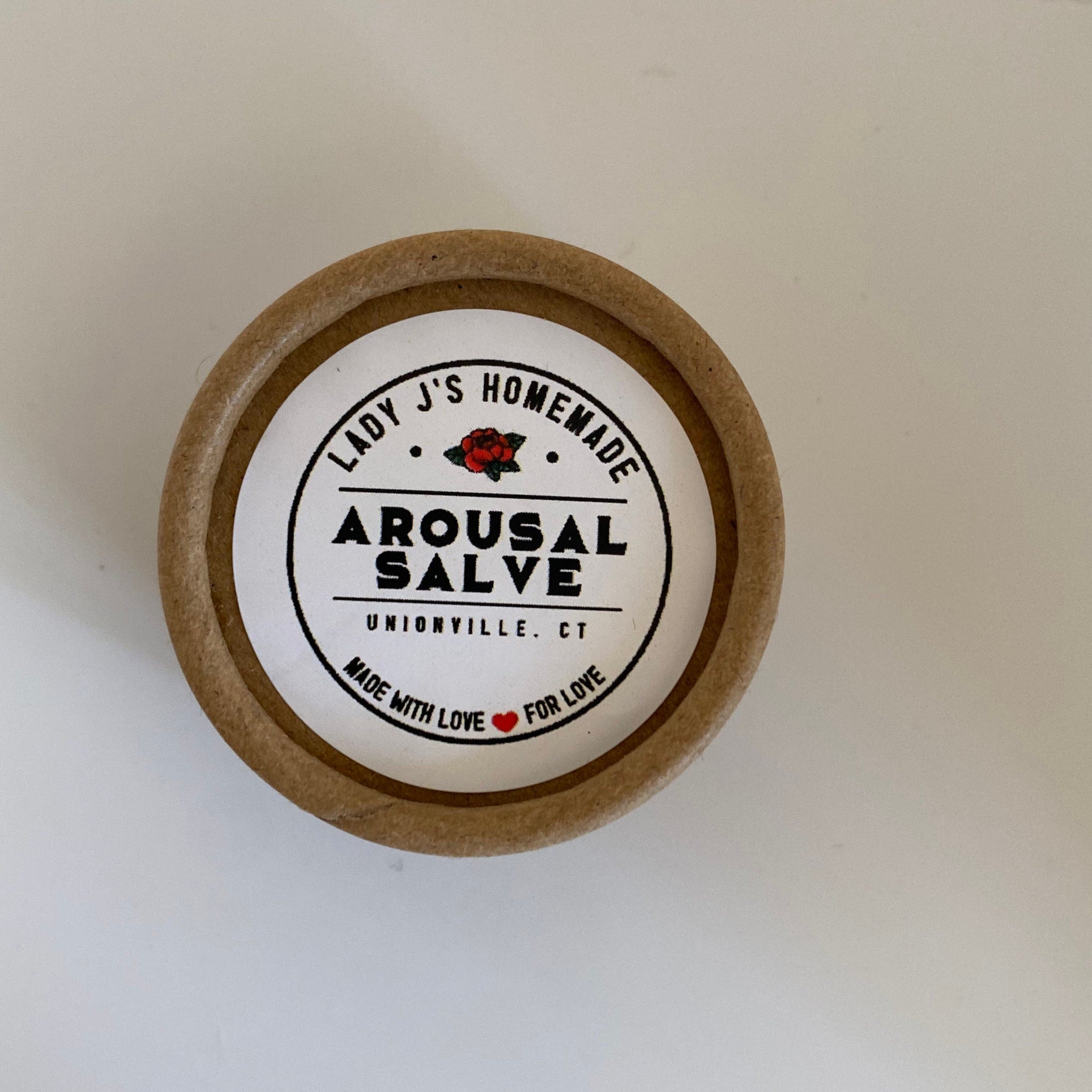 Arousal salve in a brown cardboard container with white label and black text, "Lady J's Homemade" brand name visible