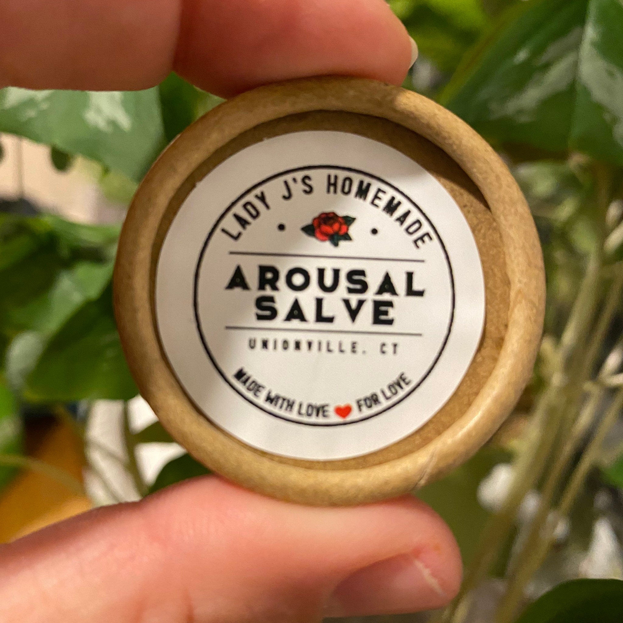 Arousal salve in round cardboard container with white label and Lady J's Homemade text, held by hand near green plants