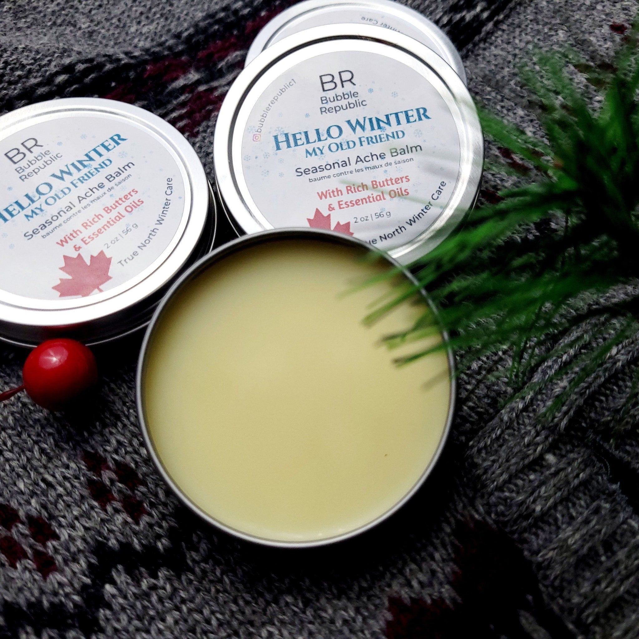 Seasonal Ache Balm tins with silver lids, yellow balm, and text from Bubble Republic on gray knit fabric