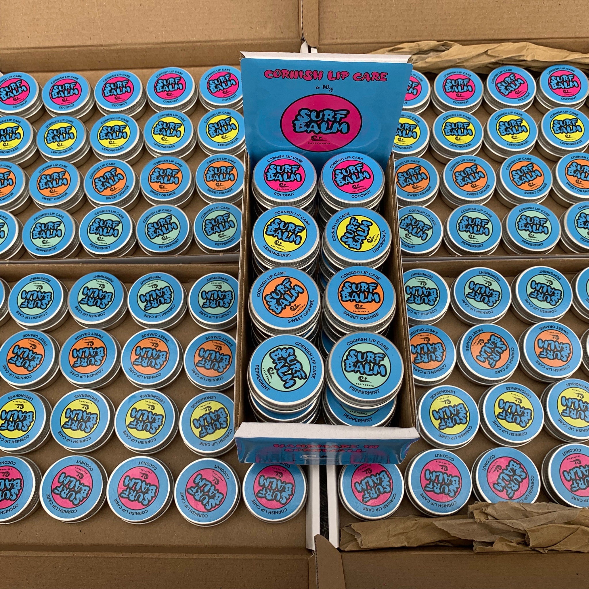 Lip balm tins with blue, pink, and orange labels, arranged in a box for display