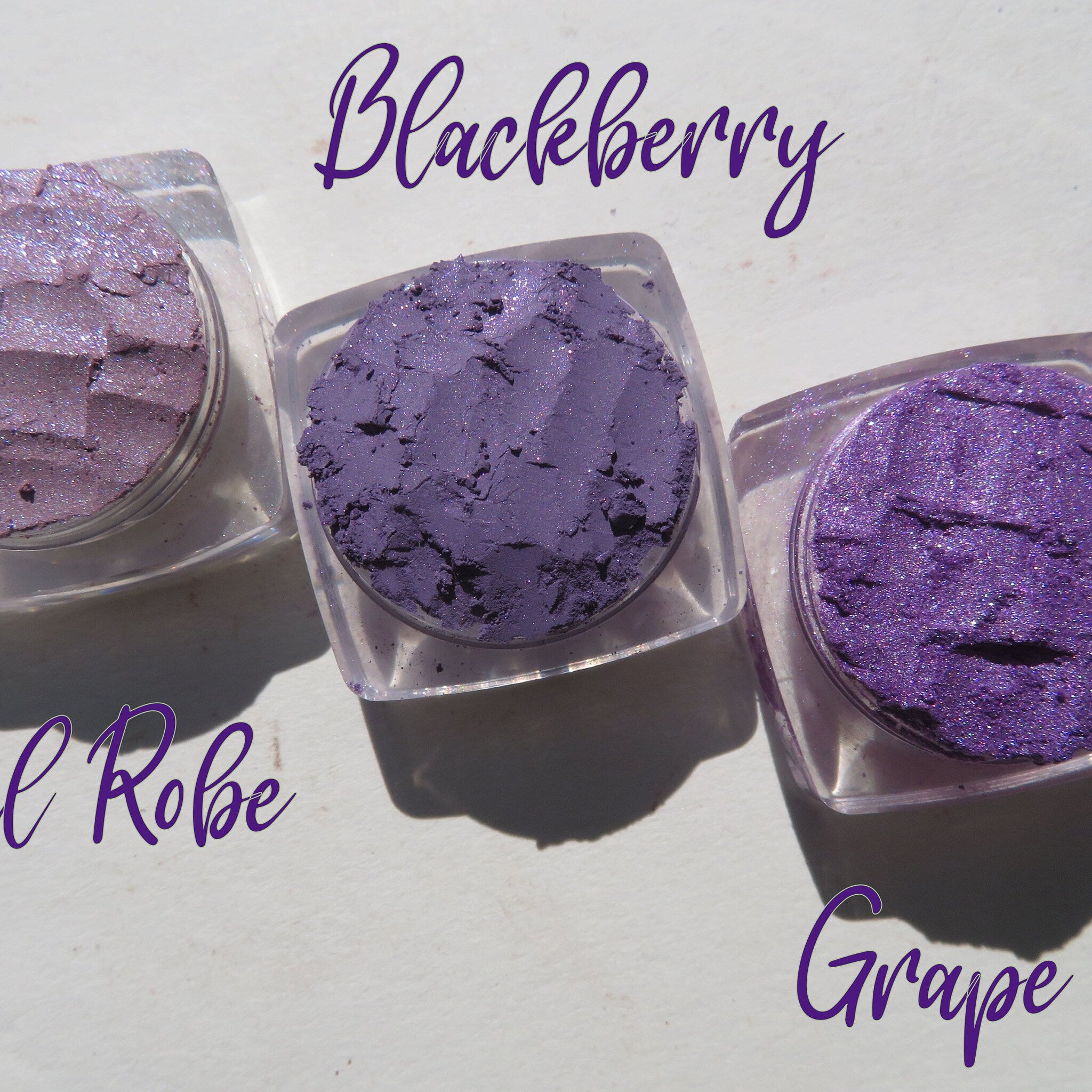 Three vibrant purple loose mineral eyeshadows with shimmer in glass jars, arranged on a sleek black tray.