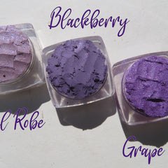Three vibrant purple loose mineral eyeshadows with shimmer in glass jars, arranged on a sleek black tray.