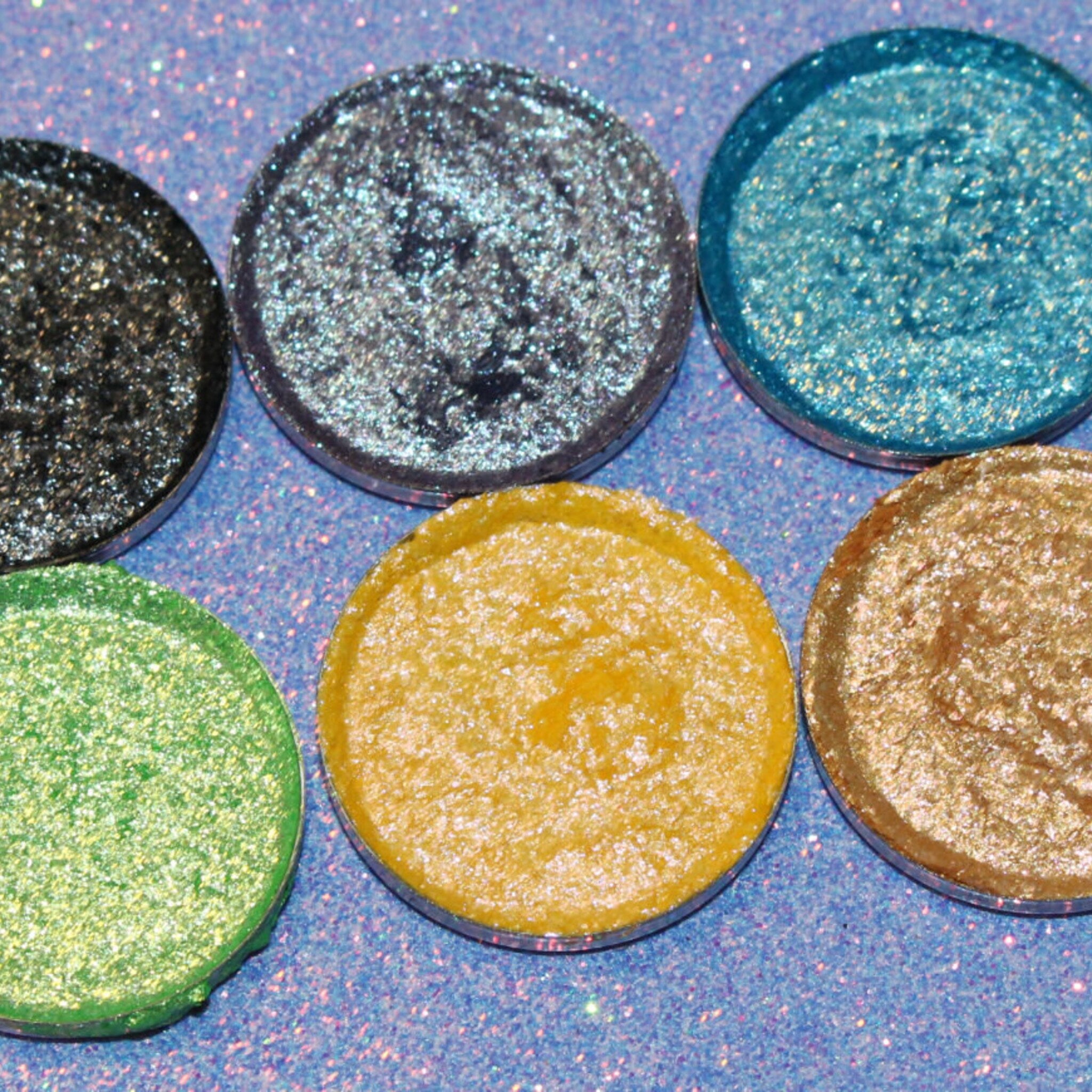 Six shimmering eyeshadow pans in bronze, gold, pink, and plum hues arranged in a sleek black palette