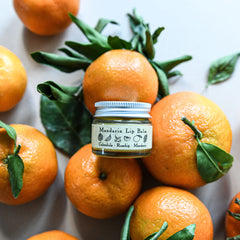 Lip balm jar with silver lid, clear glass, and "Mandarin Lip Balm" label near oranges and green leaves