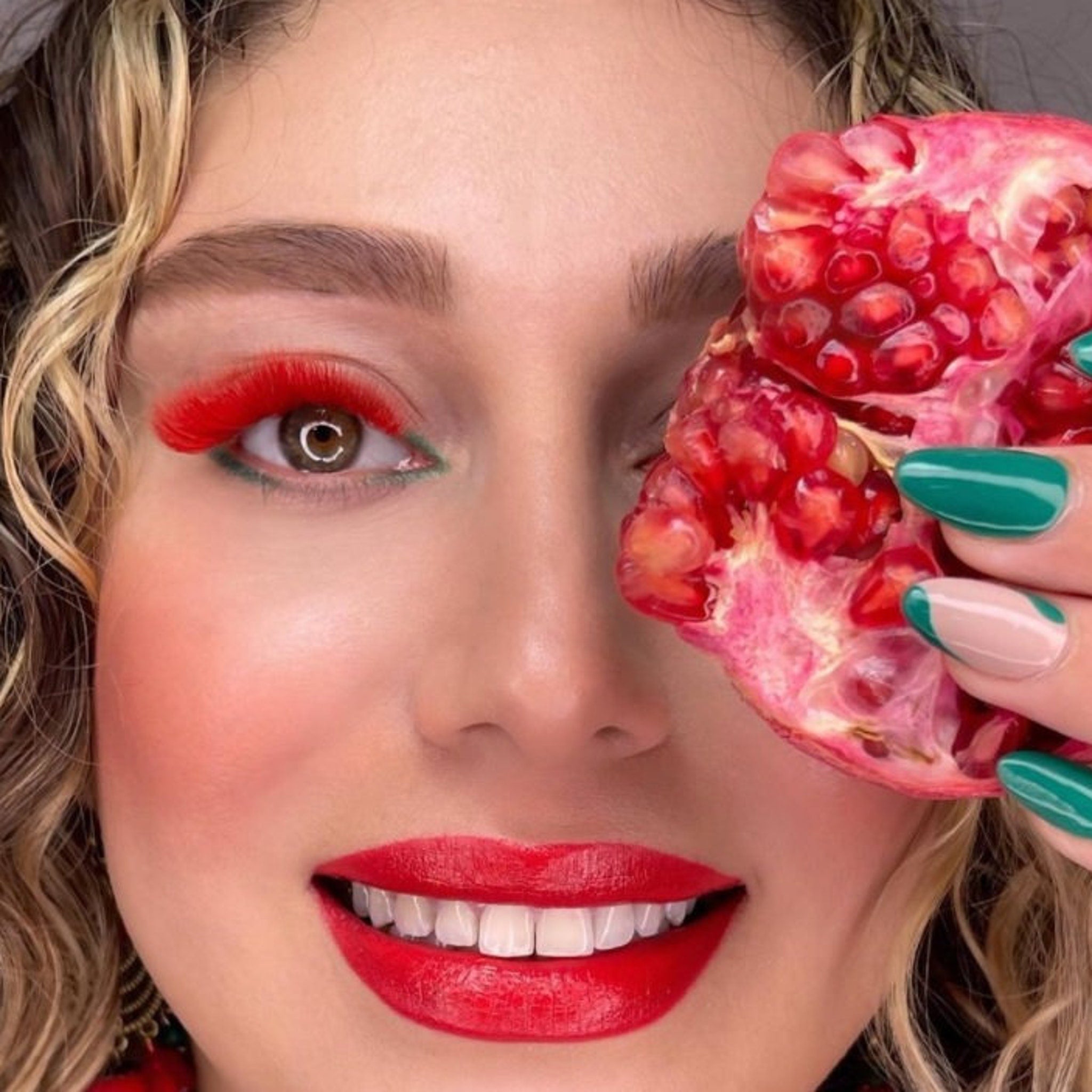 Red eyelashes with red lips and green nail polish holding pomegranate