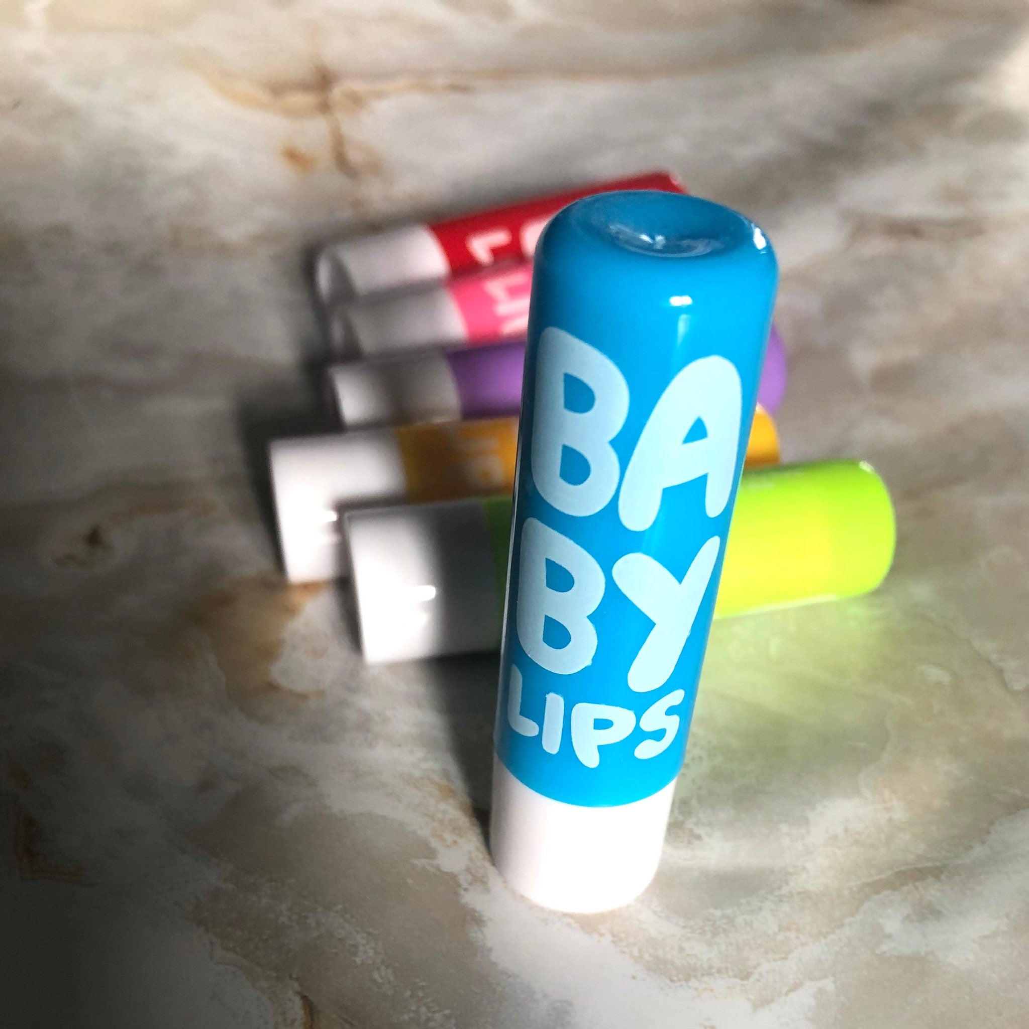 Lip balm tubes, blue and white with "BABY LIPS" text, colorful tubes in background on surface