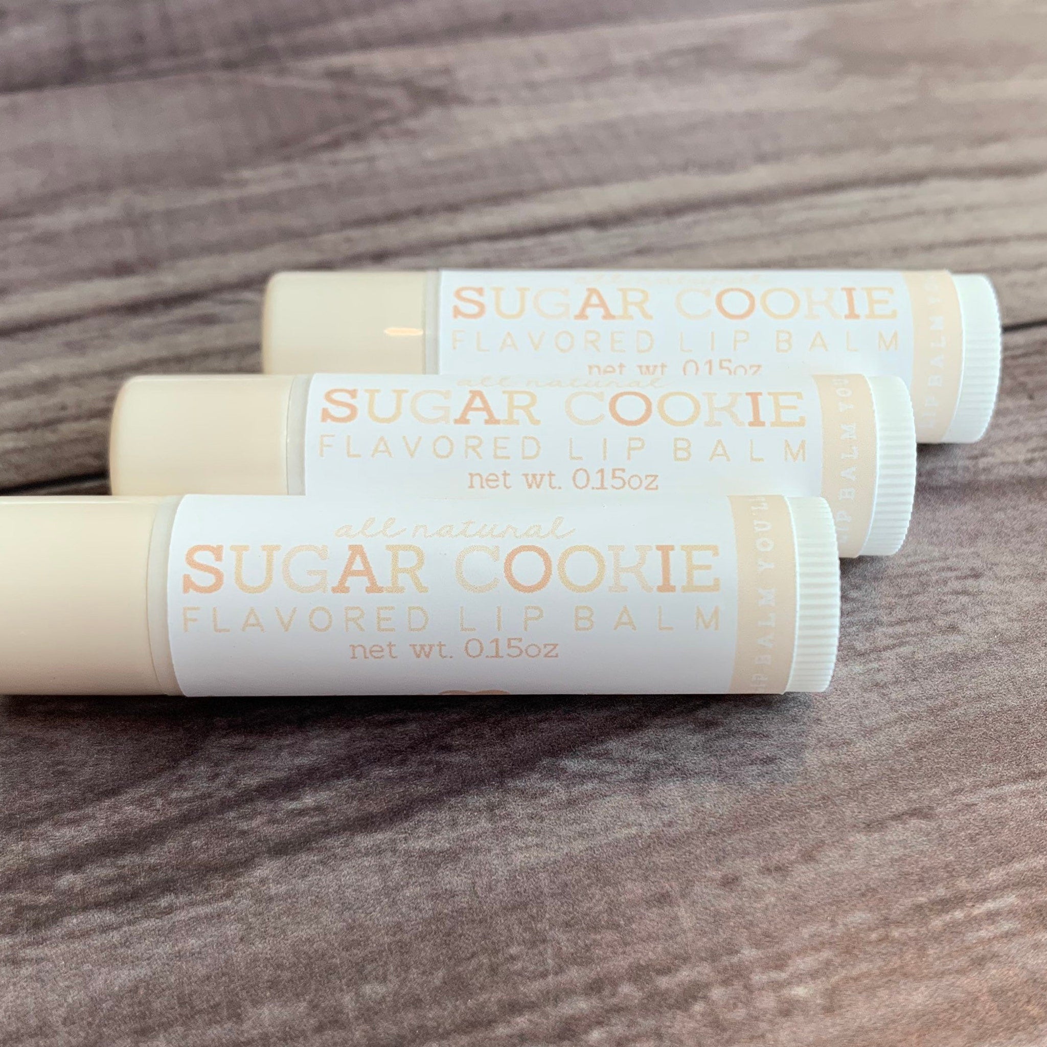 Lip balm tubes with white and peach labels, "Sugar Cookie" text, and beige tubes on wood surface