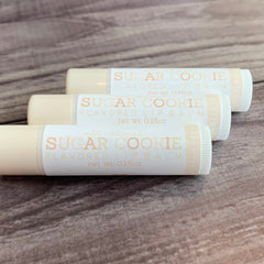 Lip balm tubes with white and peach labels, "Sugar Cookie" text, and beige tubes on wood surface