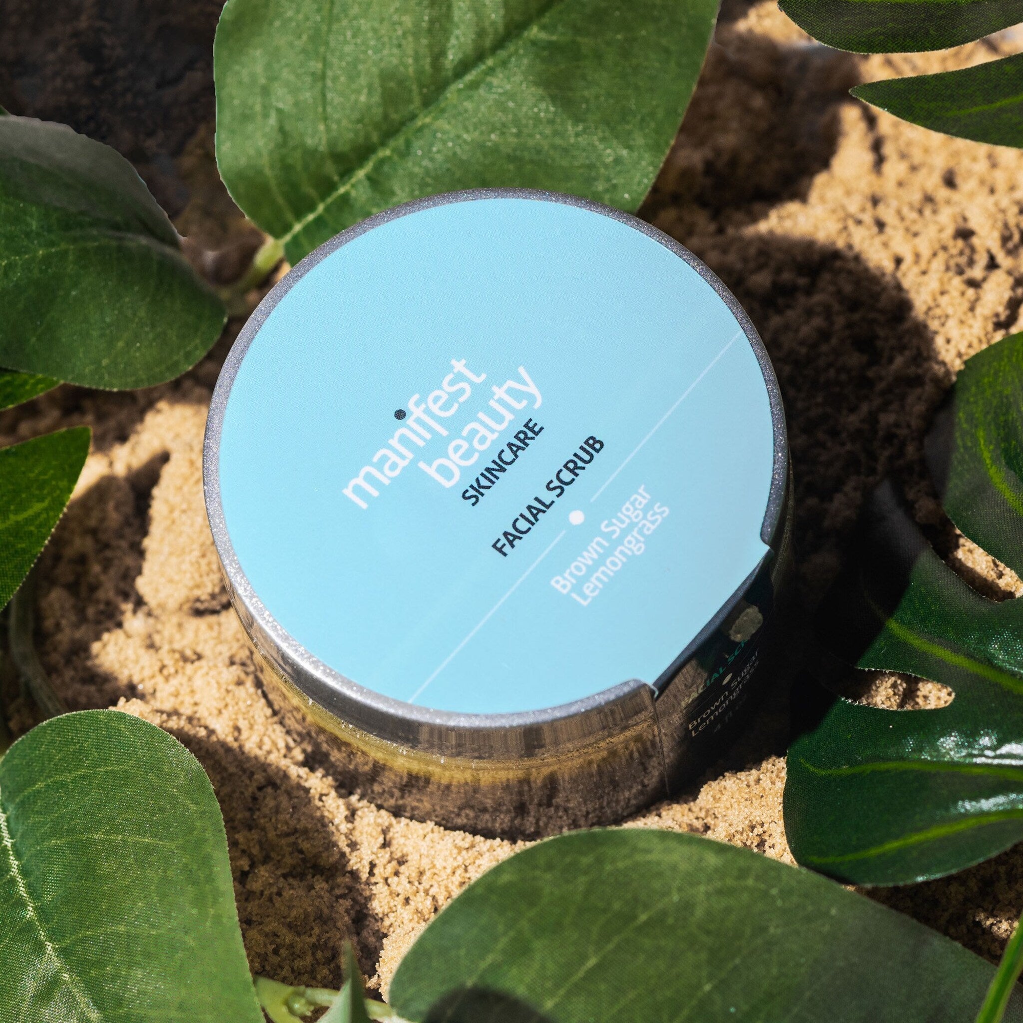 Facial scrub jar with light blue lid, silver rim, and manifest beauty label on sand and leaves