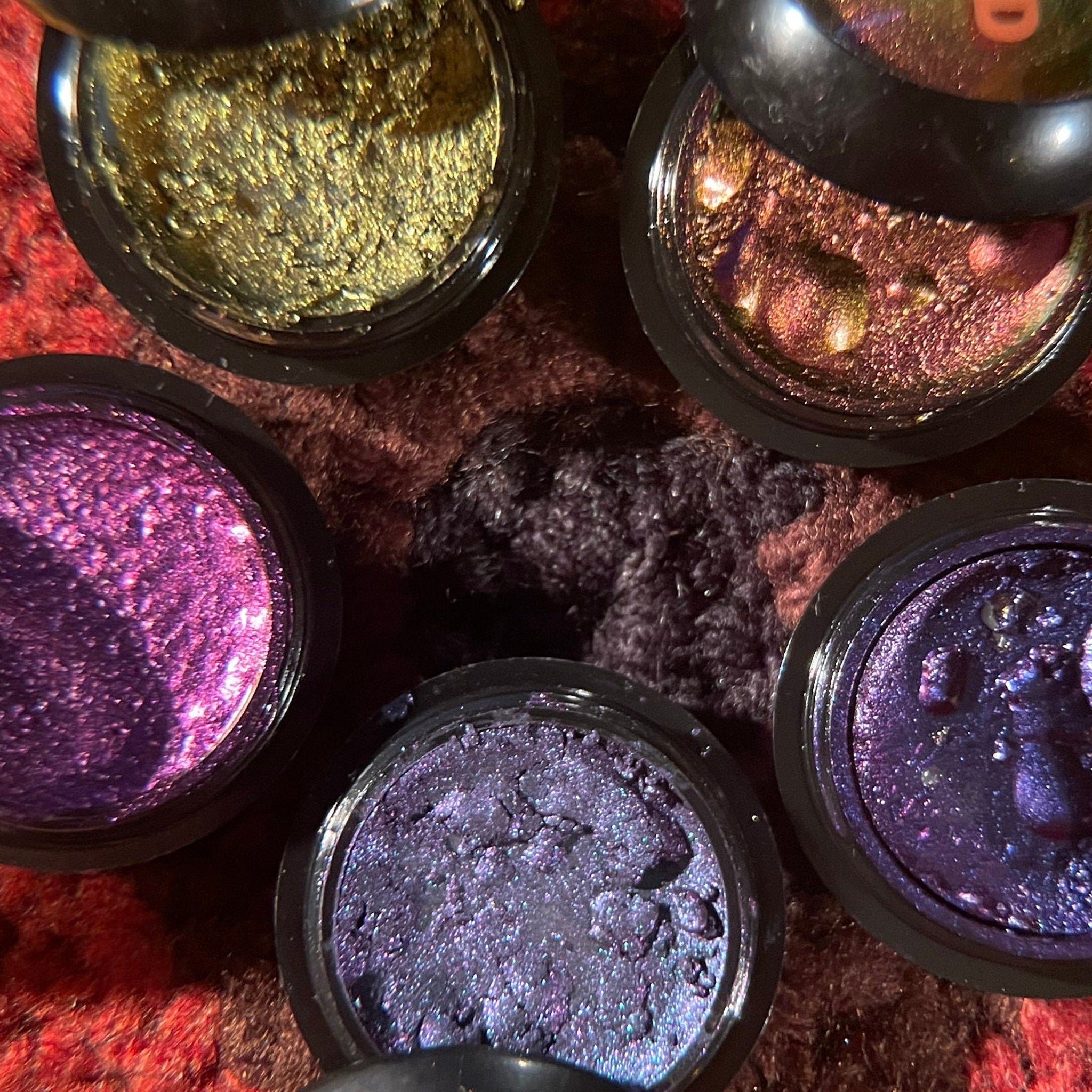 Eyeshadow pots with metallic gold, purple, and blue shimmer, in black containers, on textured fabric
